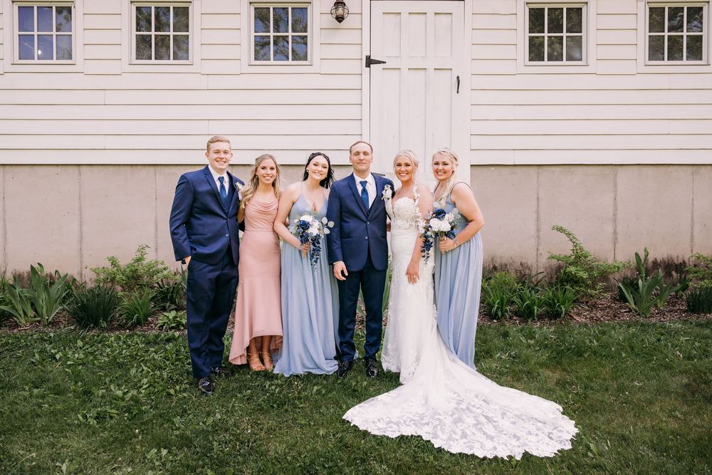 Wedding party posing outdoors, bride in lace gown with bouquet, bridesmaids in blue and pink dresses, groomsmen in navy suits.
