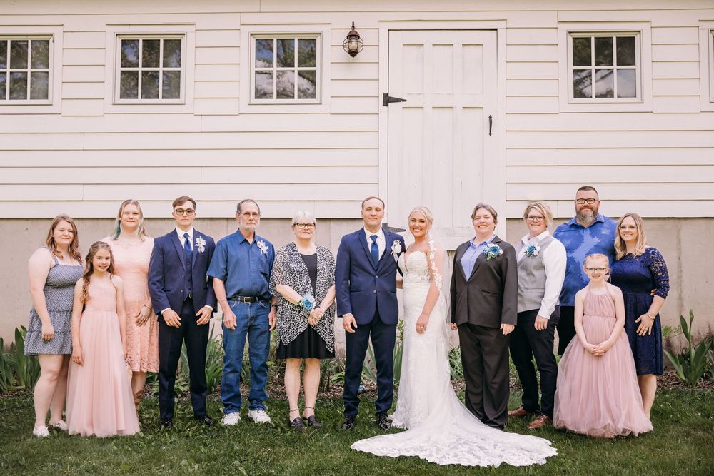 Wedding party poses outdoors in front of white barn, bride in white gown with groom and family in formal attire.