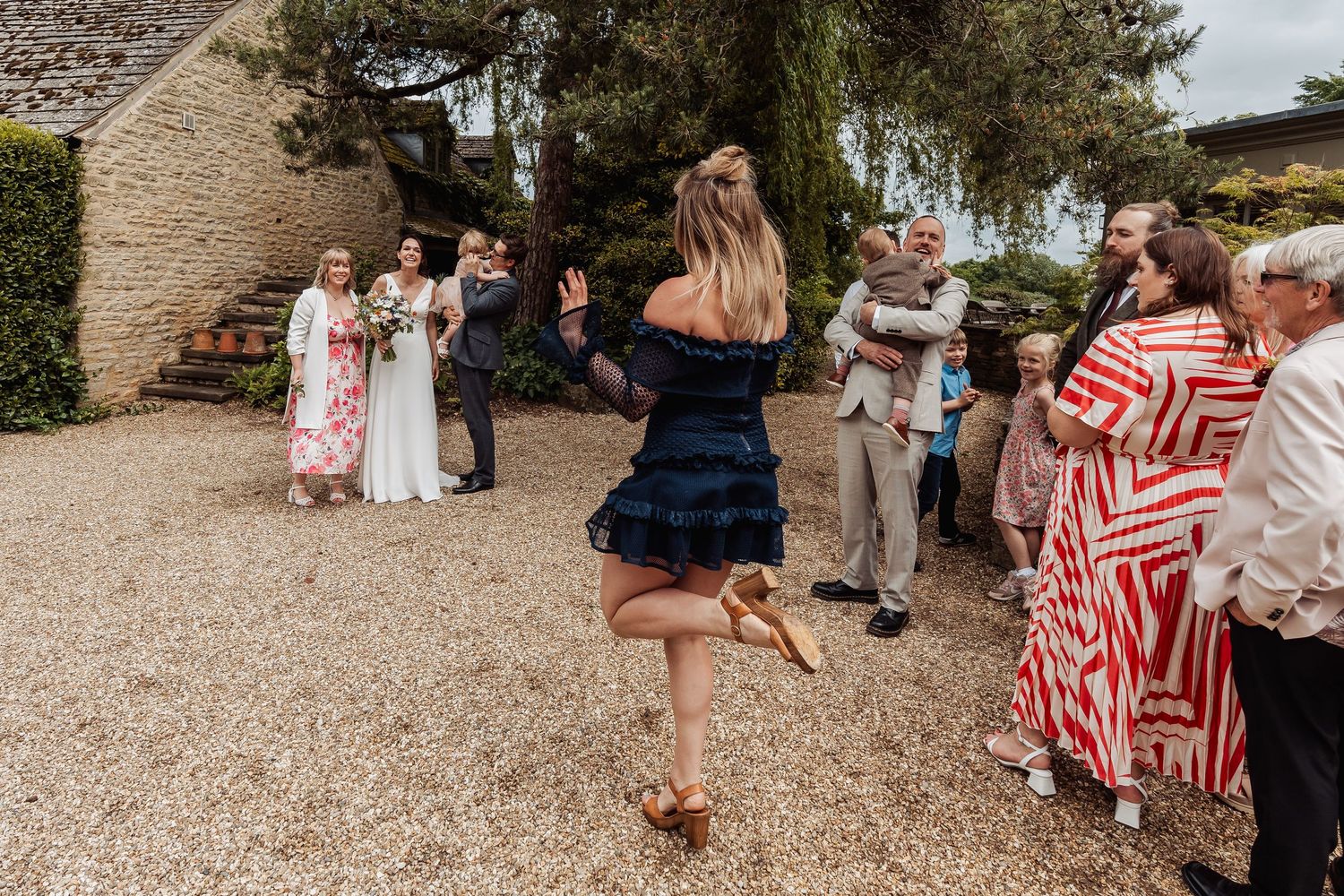Wedding guests mingle outdoors as a woman in navy dress dances toward a small child at a rustic venue.