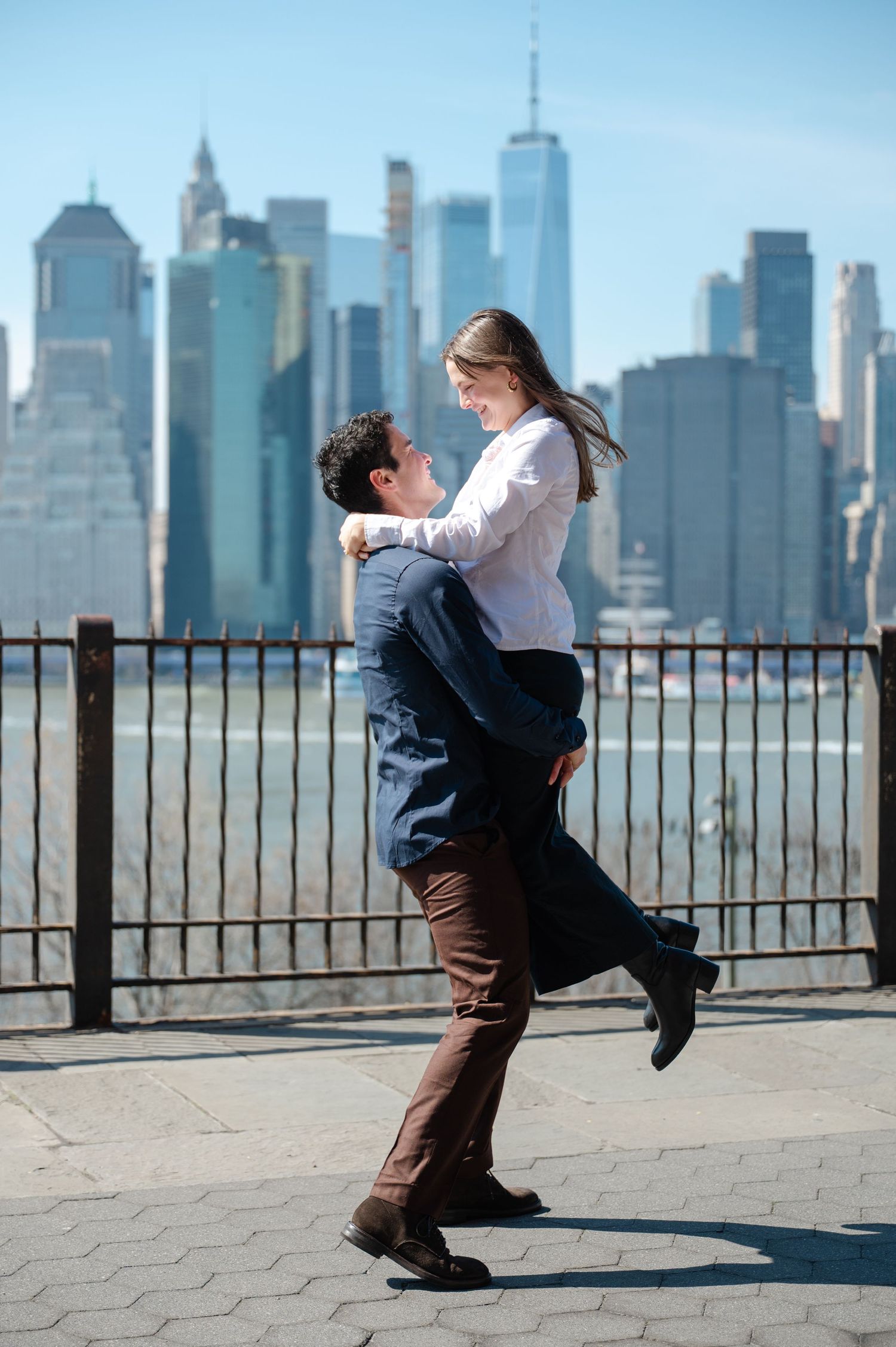 Couple embracing with NYC skyline backdrop, man lifting woman joyfully on Brooklyn waterfront promenade after a proposal