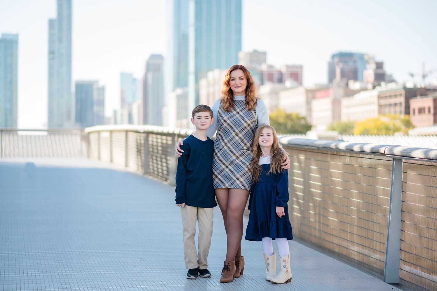 A mother poses with her three young sons on a waterfront bridge in an urban park setting on a sunny day.