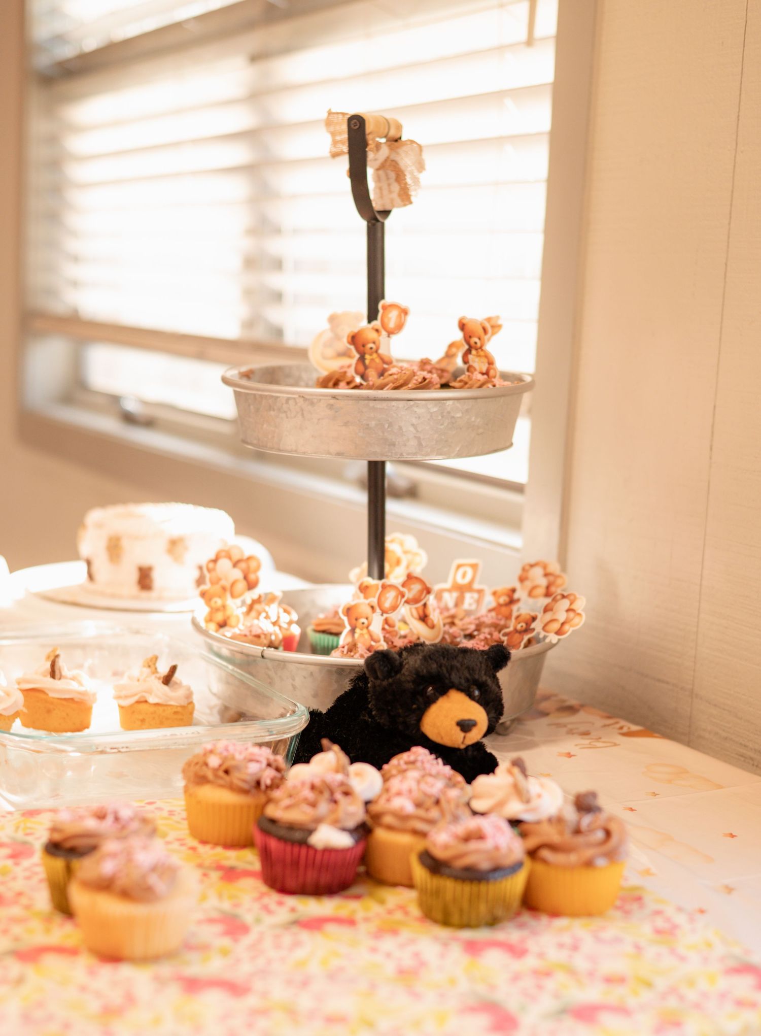A tiered metal stand with bear-shaped treats and cake pops, surrounded by frosted cupcakes and a black bear plush on a floral tablecloth.