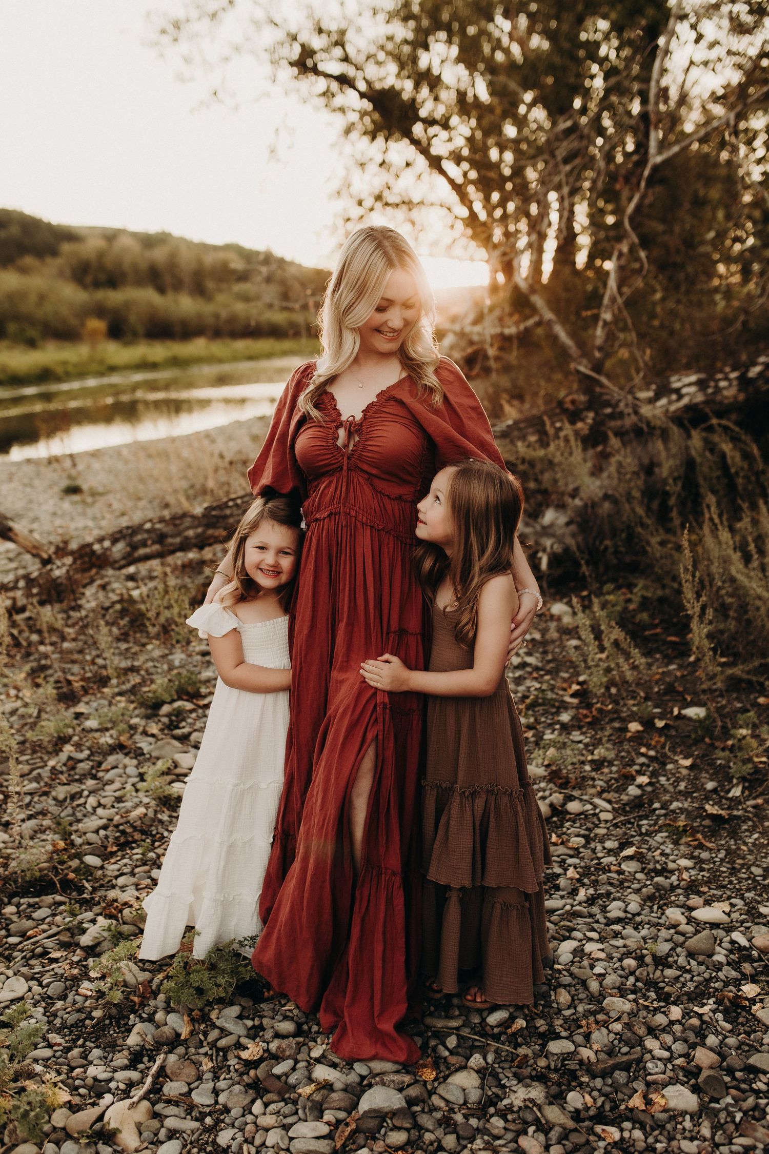 A pregnant woman in a red boho dress poses outdoors with two young daughters at golden hour near a rocky riverbank.