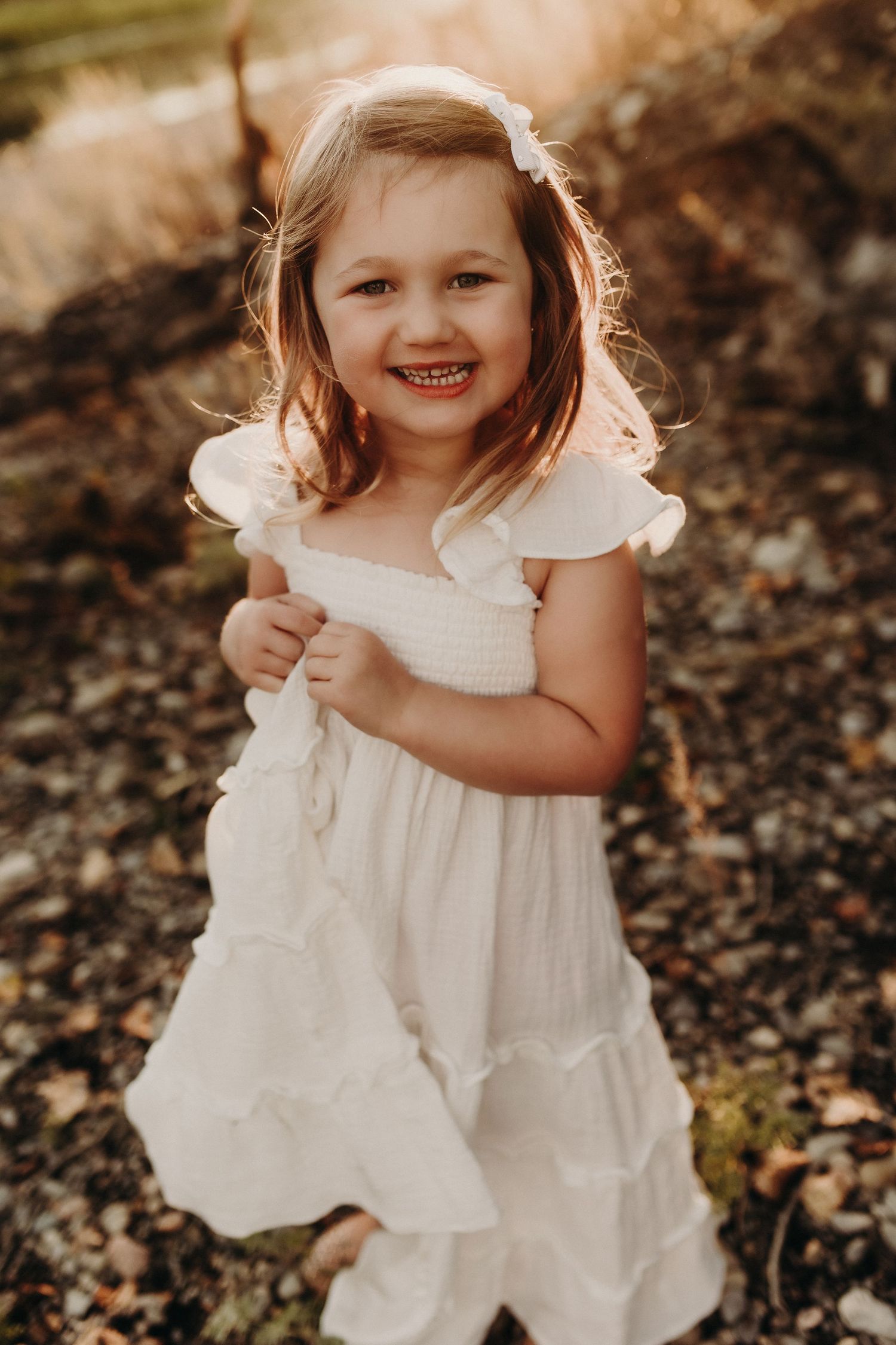 Happy young girl in white ruffled dress smiles outdoors in golden sunlight.