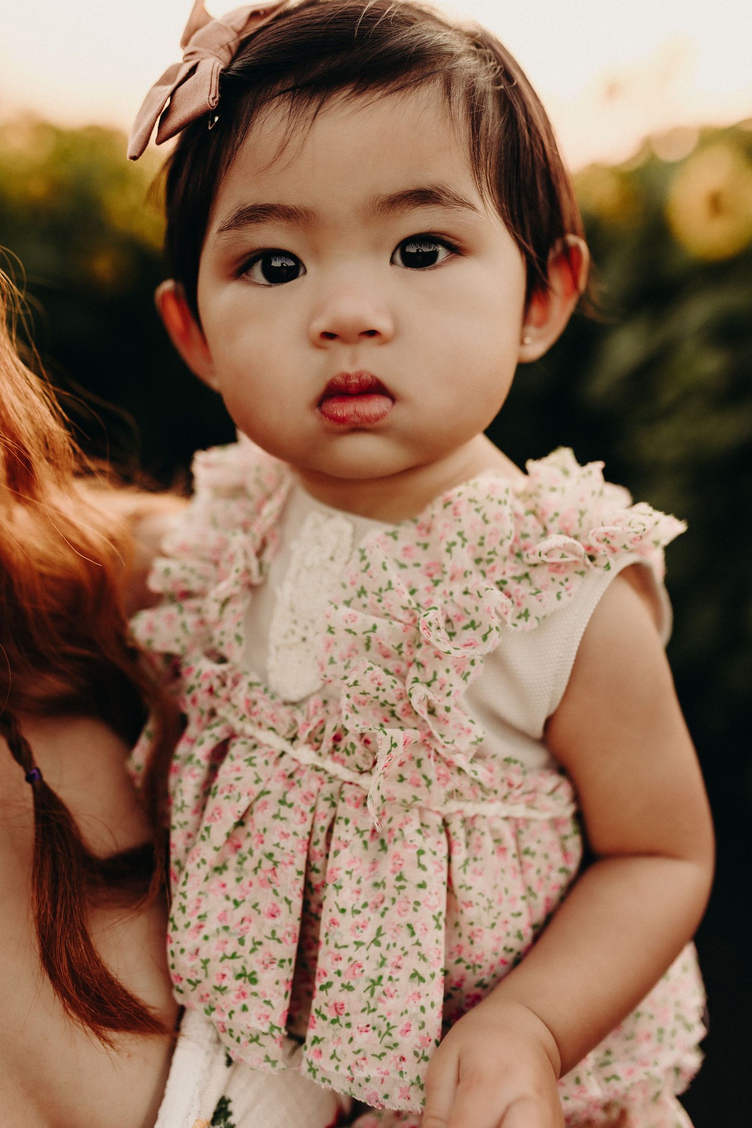 Adorable baby girl in floral ruffle dress with bow headband, held outdoors in golden hour light.