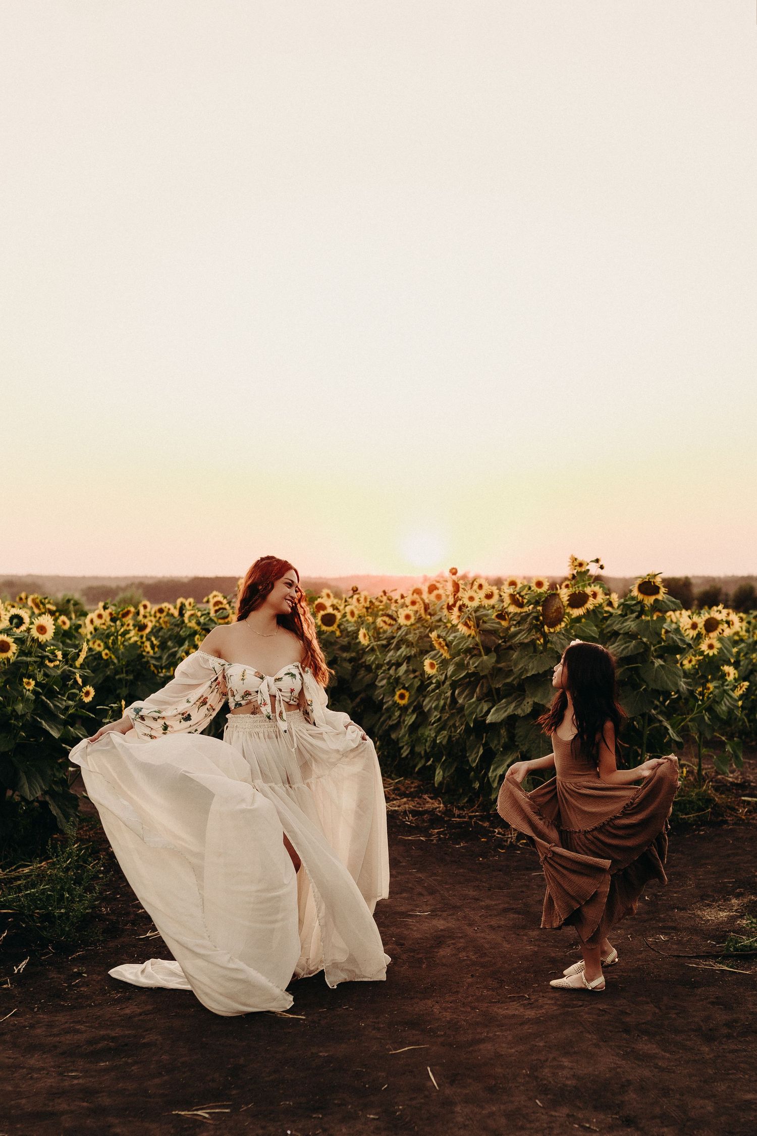 Two women in flowing dresses pose in a sunflower field at golden sunset, one in white floral gown, other in brown dress.