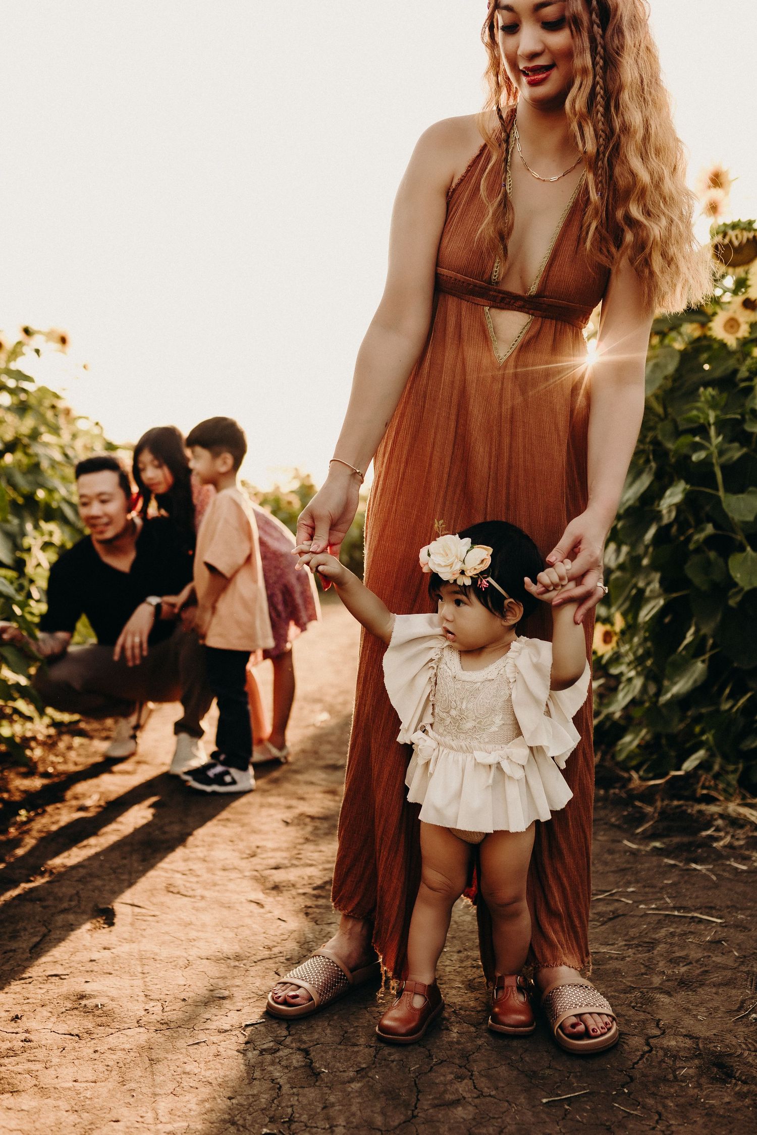 A mother in a rust dress helps her toddler walk in a sunflower field at golden hour.