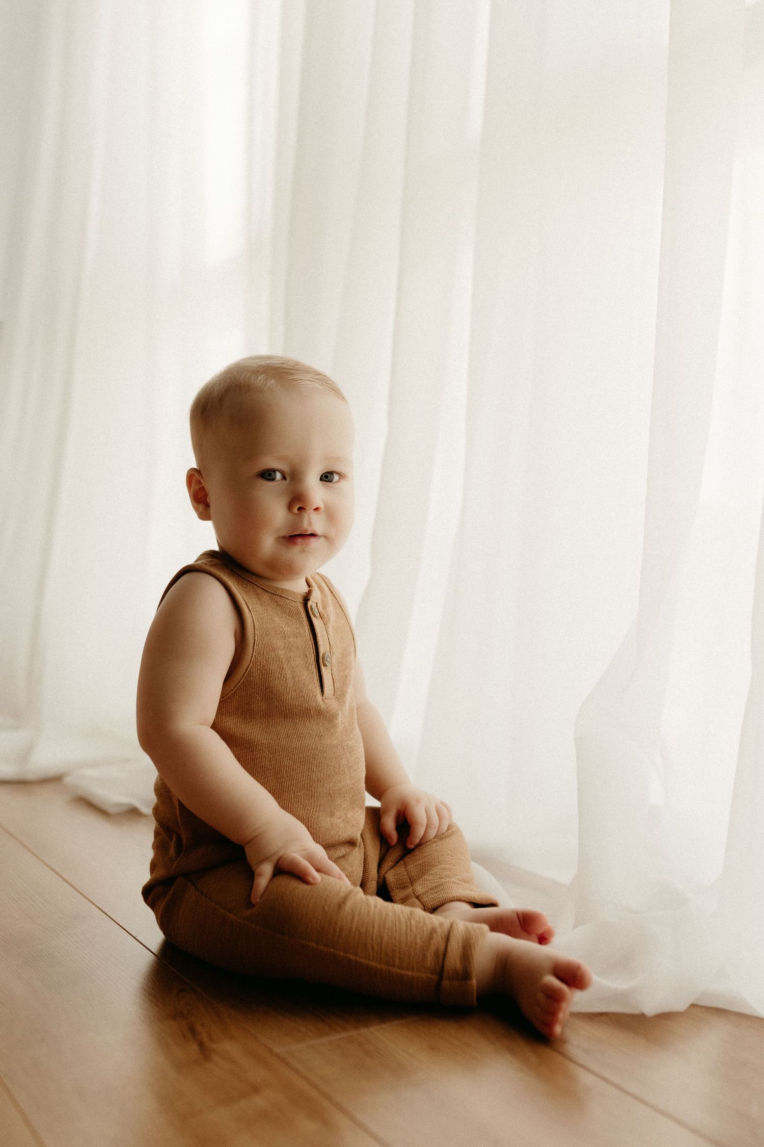 Adorable baby boy in tan linen romper sitting on hardwood floor near white curtains.