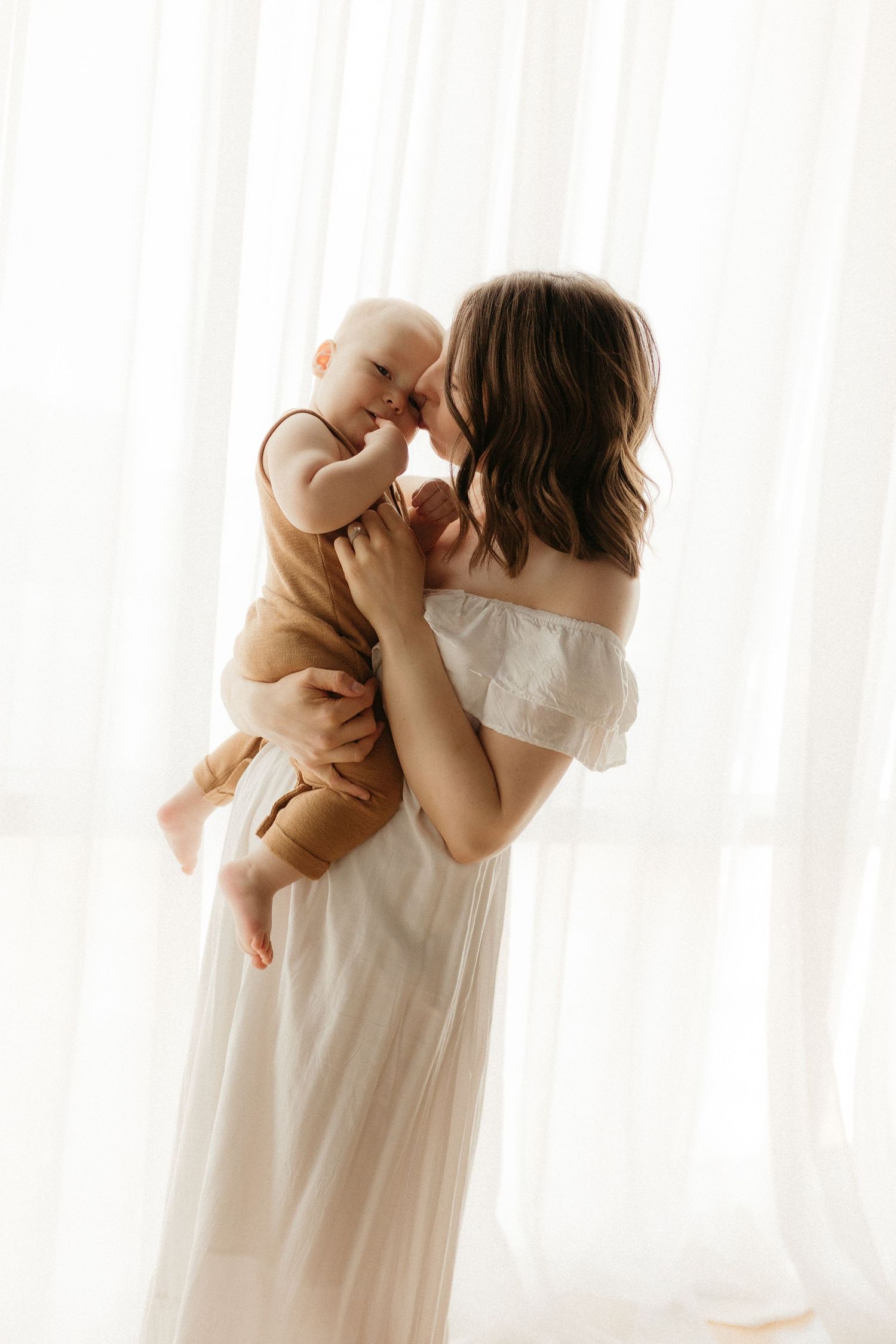 A mother in a white dress tenderly holds her smiling baby near a bright window.