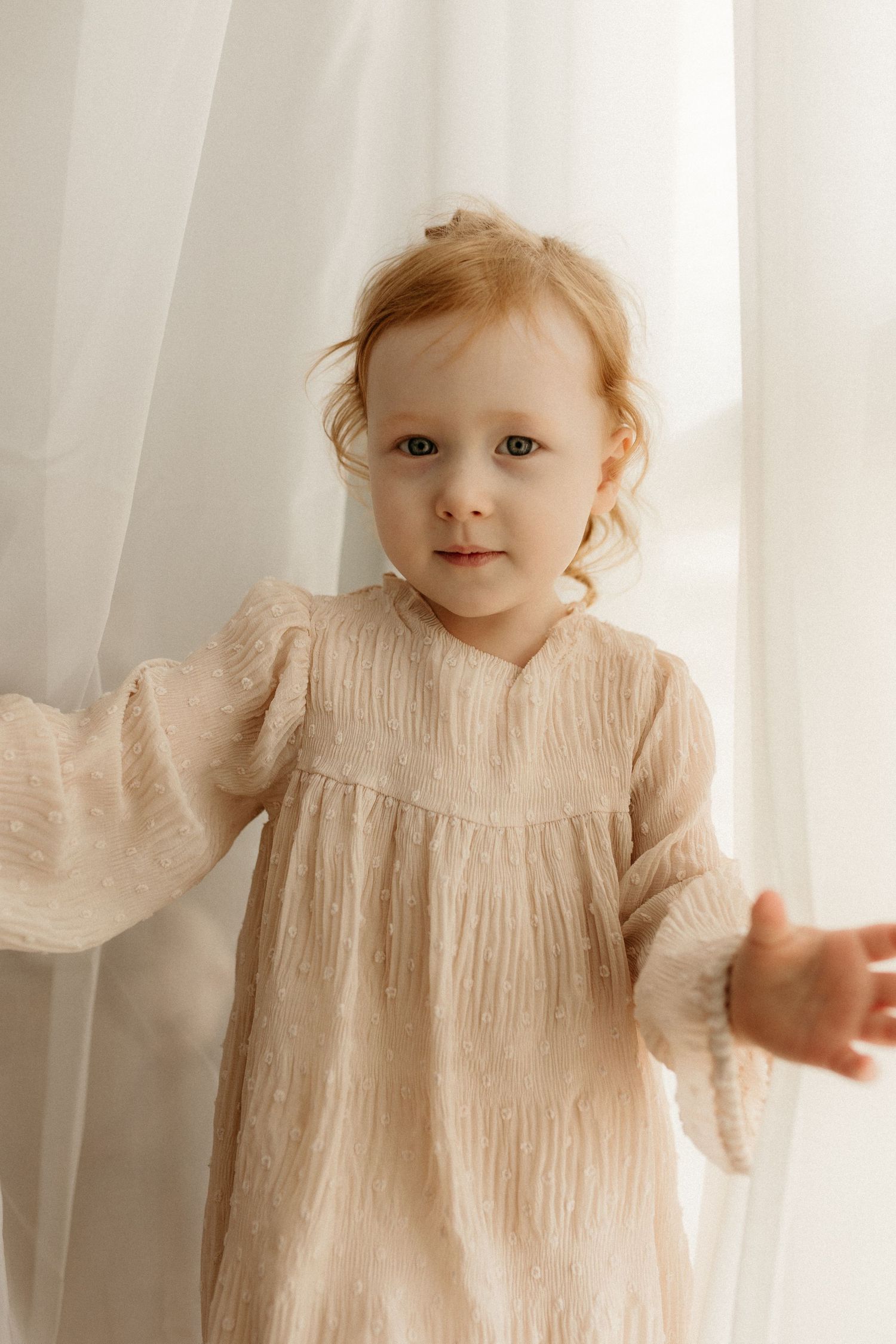 Toddler girl in cream textured dress standing by sheer white curtains, looking at camera.