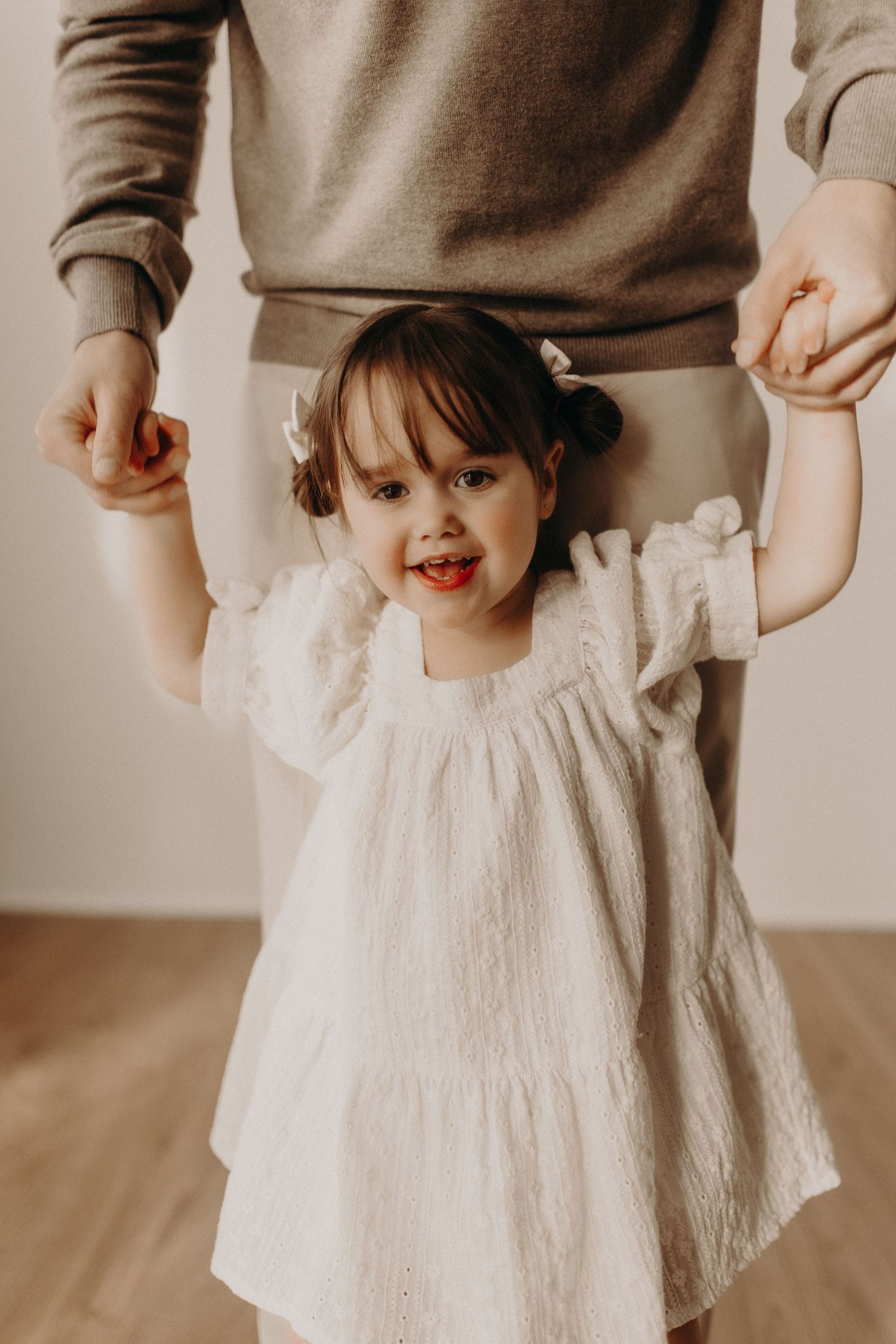 Smiling toddler girl in white ruffle dress holds adult hands while standing on hardwood floor.
