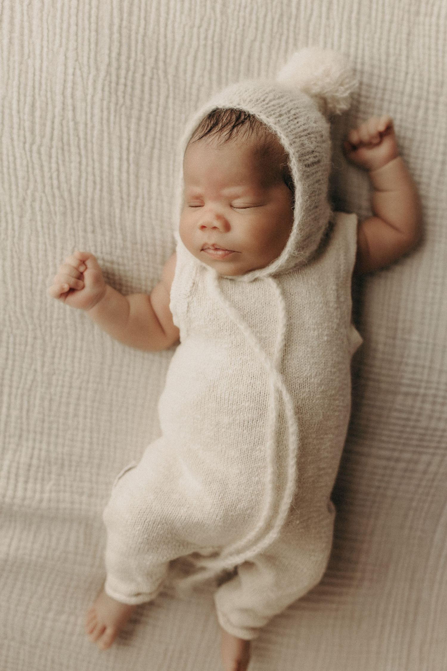 Sleeping newborn baby in white knit outfit and pom-pom bonnet lying on soft textured blanket.