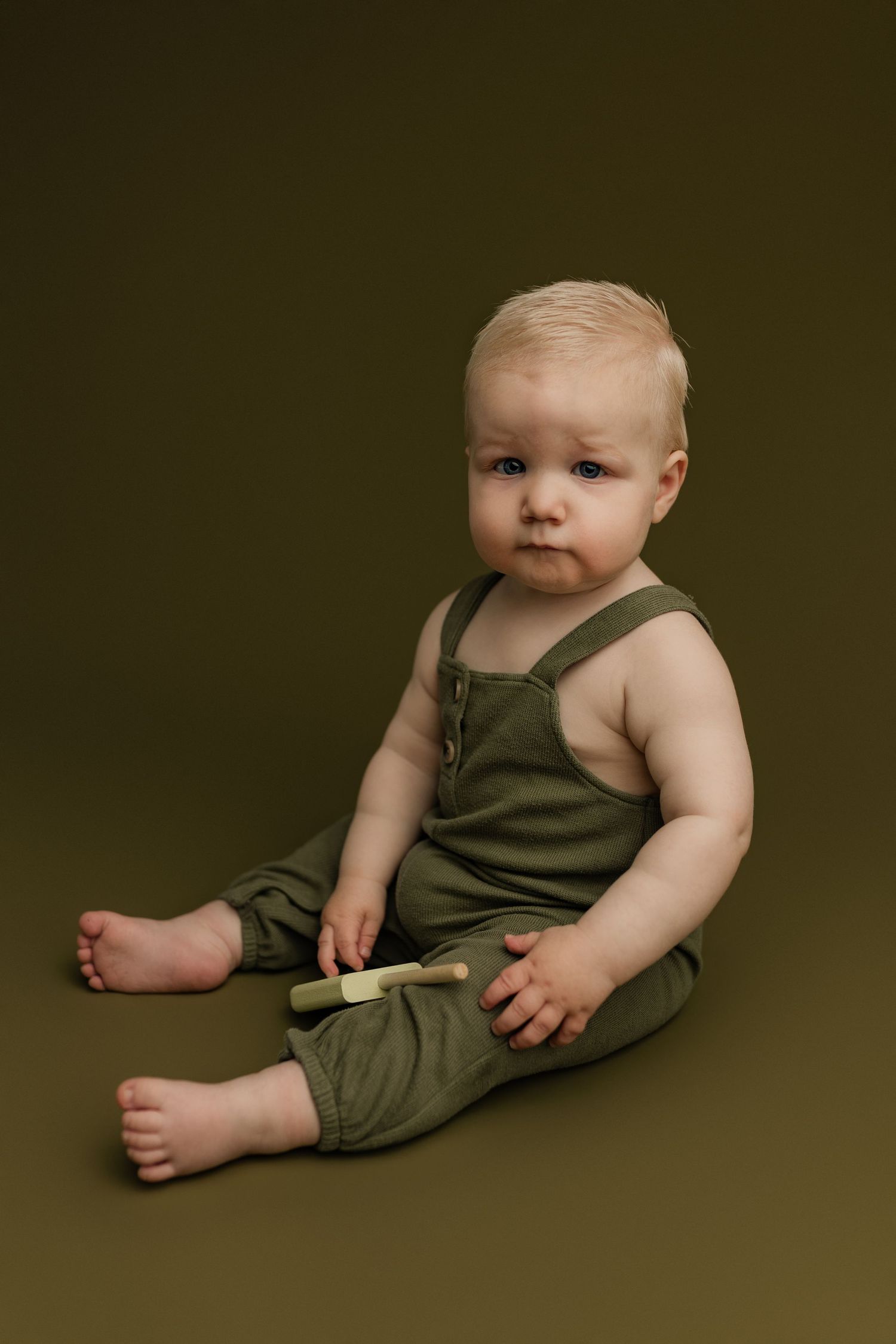Adorable blonde baby in green linen overalls sitting barefoot on olive background holding a small wooden toy.
