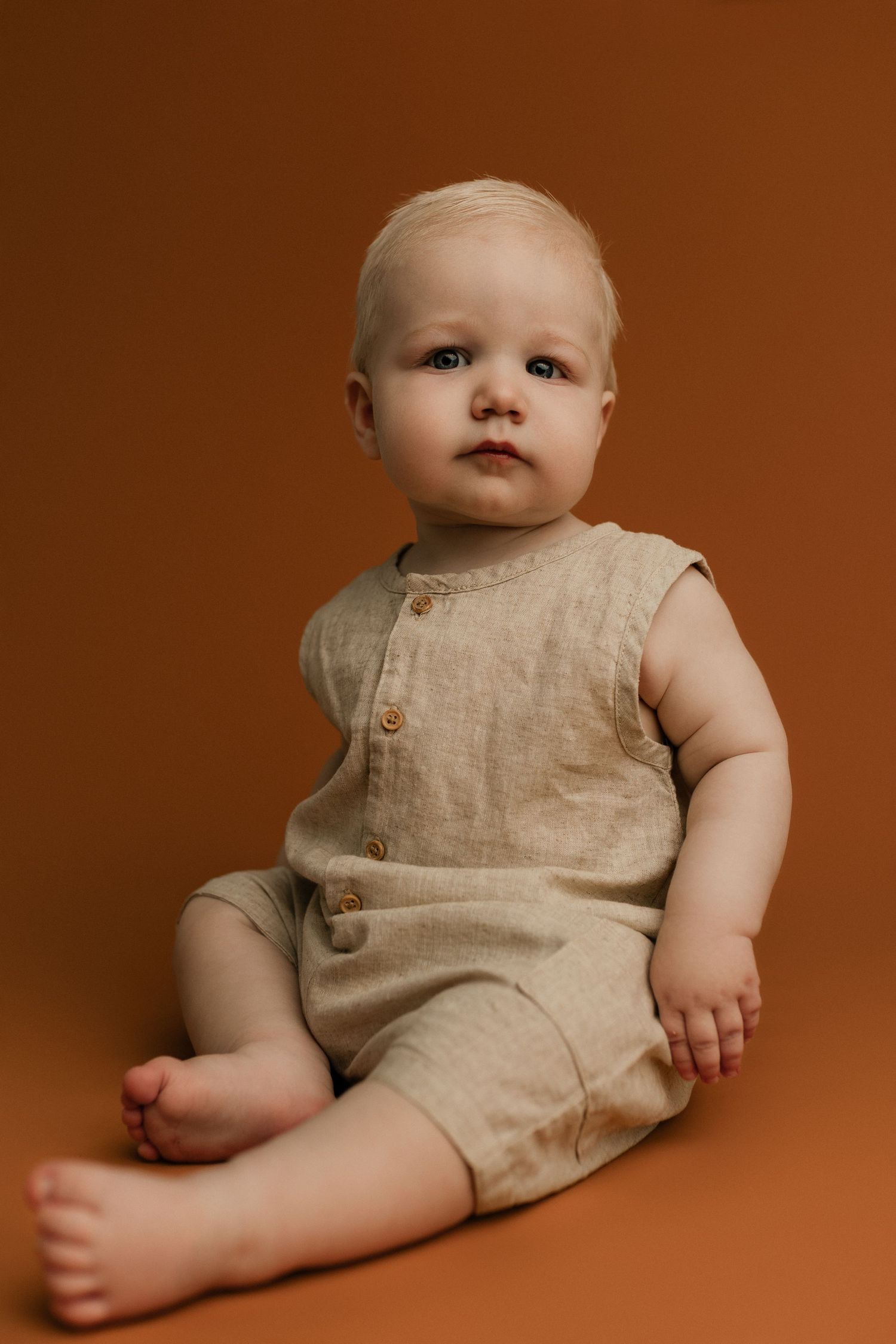 Cute baby in beige linen romper sitting on brown background, looking at camera with blue eyes.