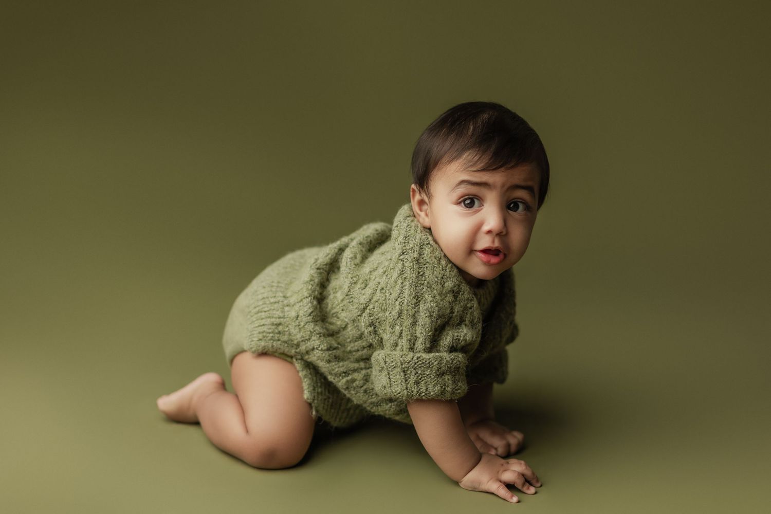 A baby in a green knit romper crawls on a green background, looking at the camera with a curious expression.