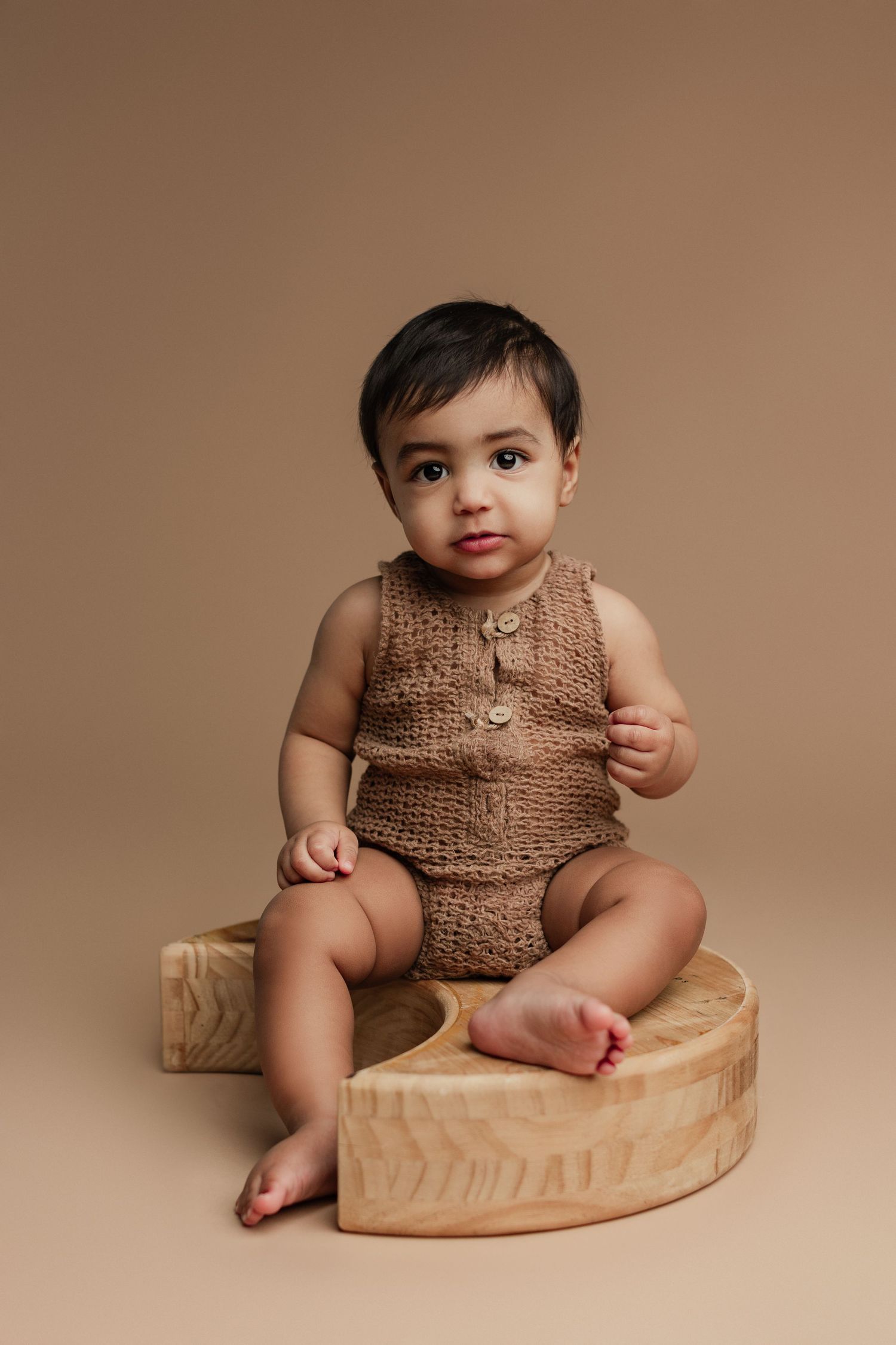 Adorable baby in brown knit romper sitting on wooden heart prop against beige background.