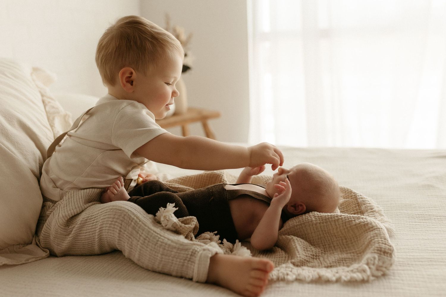 A curious toddler sits on a white bed reading a book, wrapped in a cozy knit blanket in a bright room.