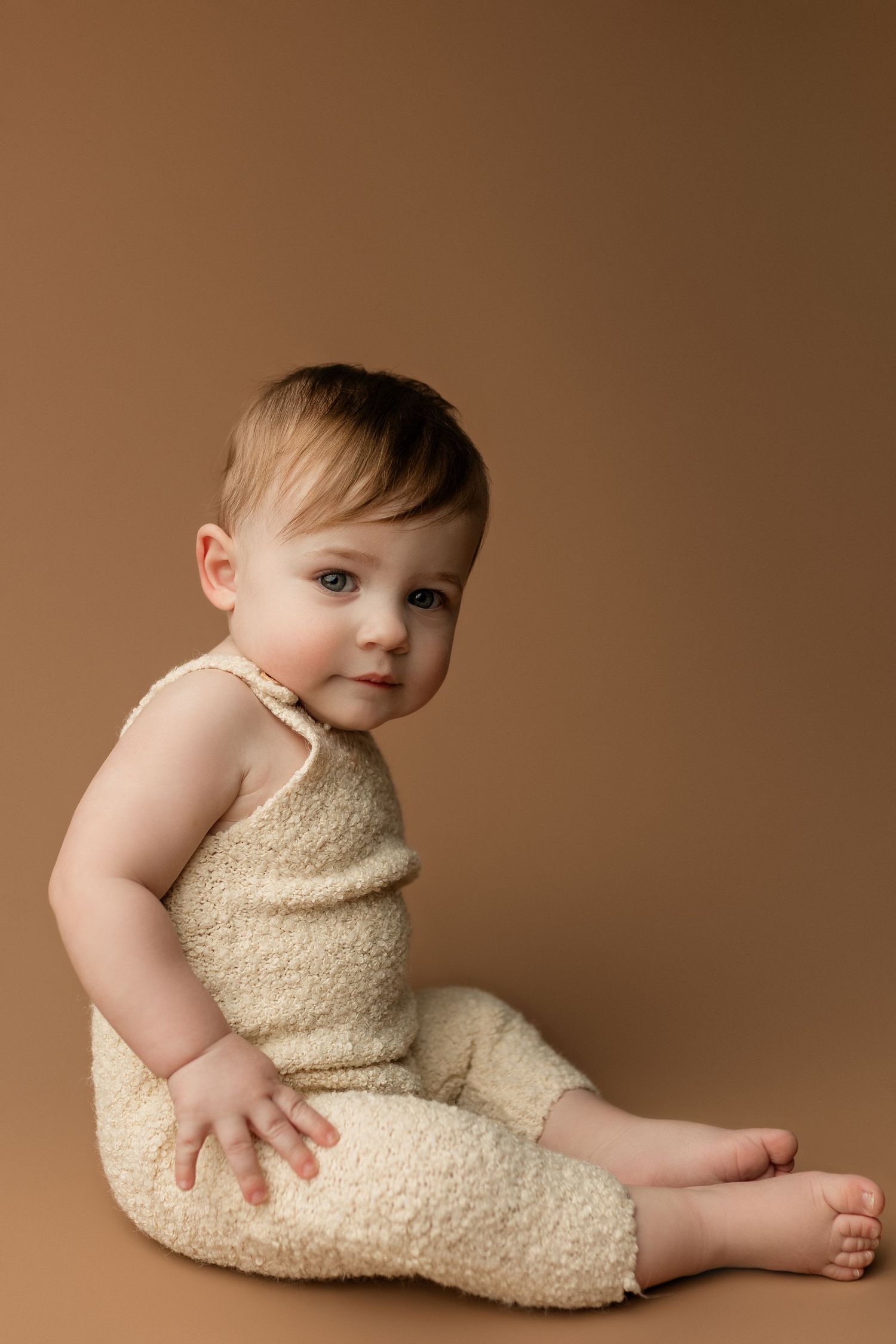 Adorable baby in cream fuzzy romper sitting on brown background, looking at camera with curious expression.