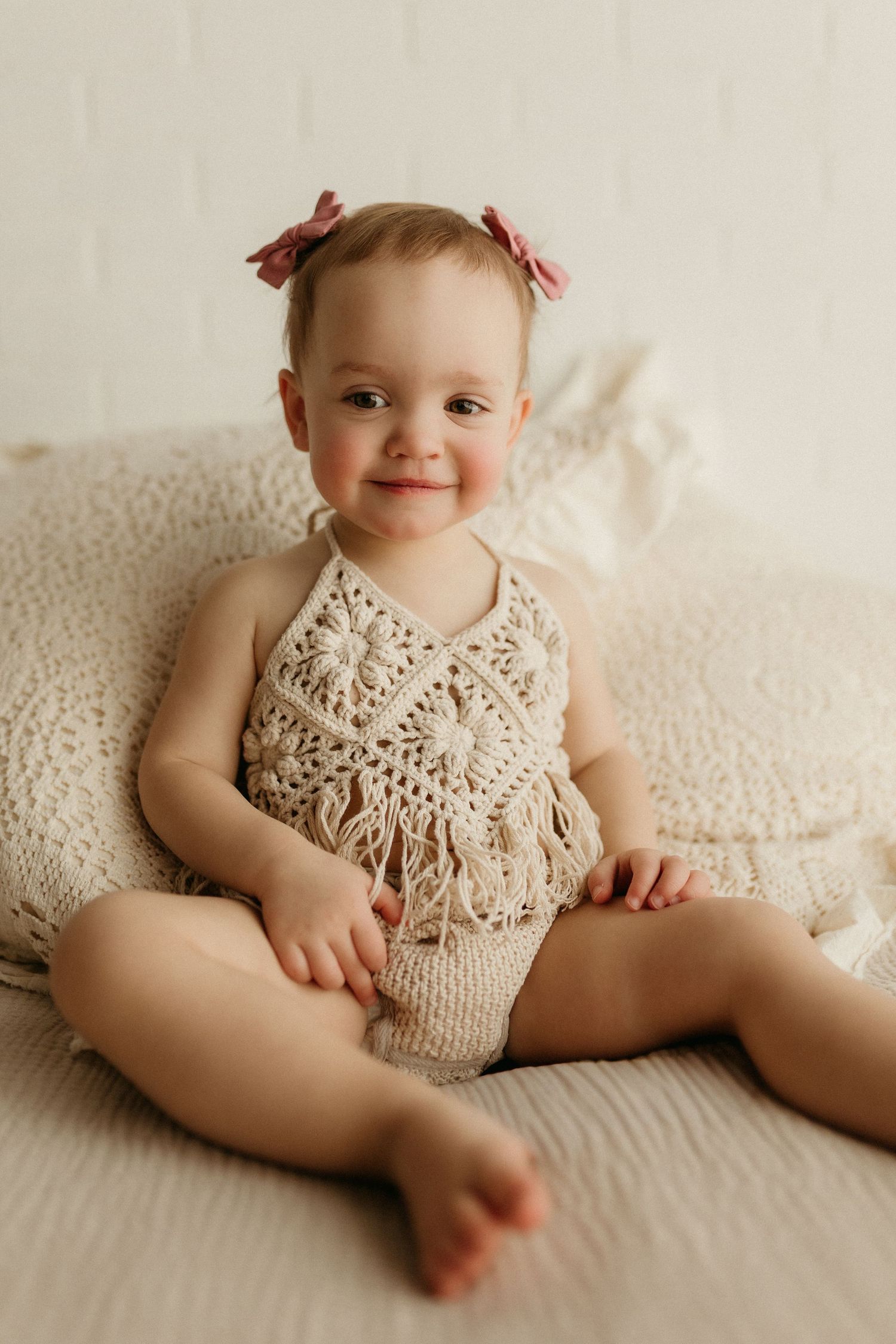 Smiling baby girl in crochet romper with pink bows sits on cream bedding for a milestone photo session.