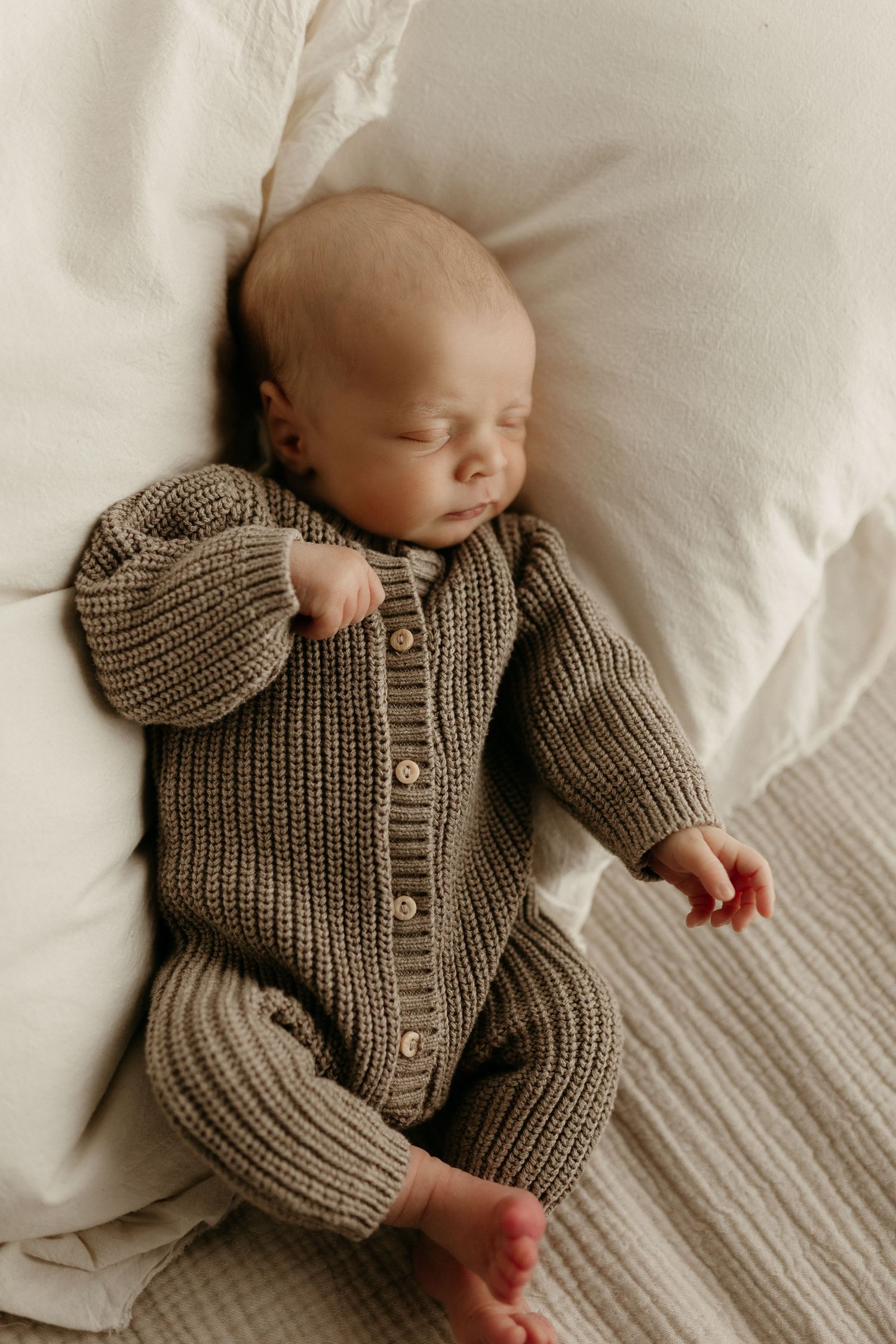Sleeping newborn baby wearing a chunky knit olive green button-up romper, lying on white bedding.