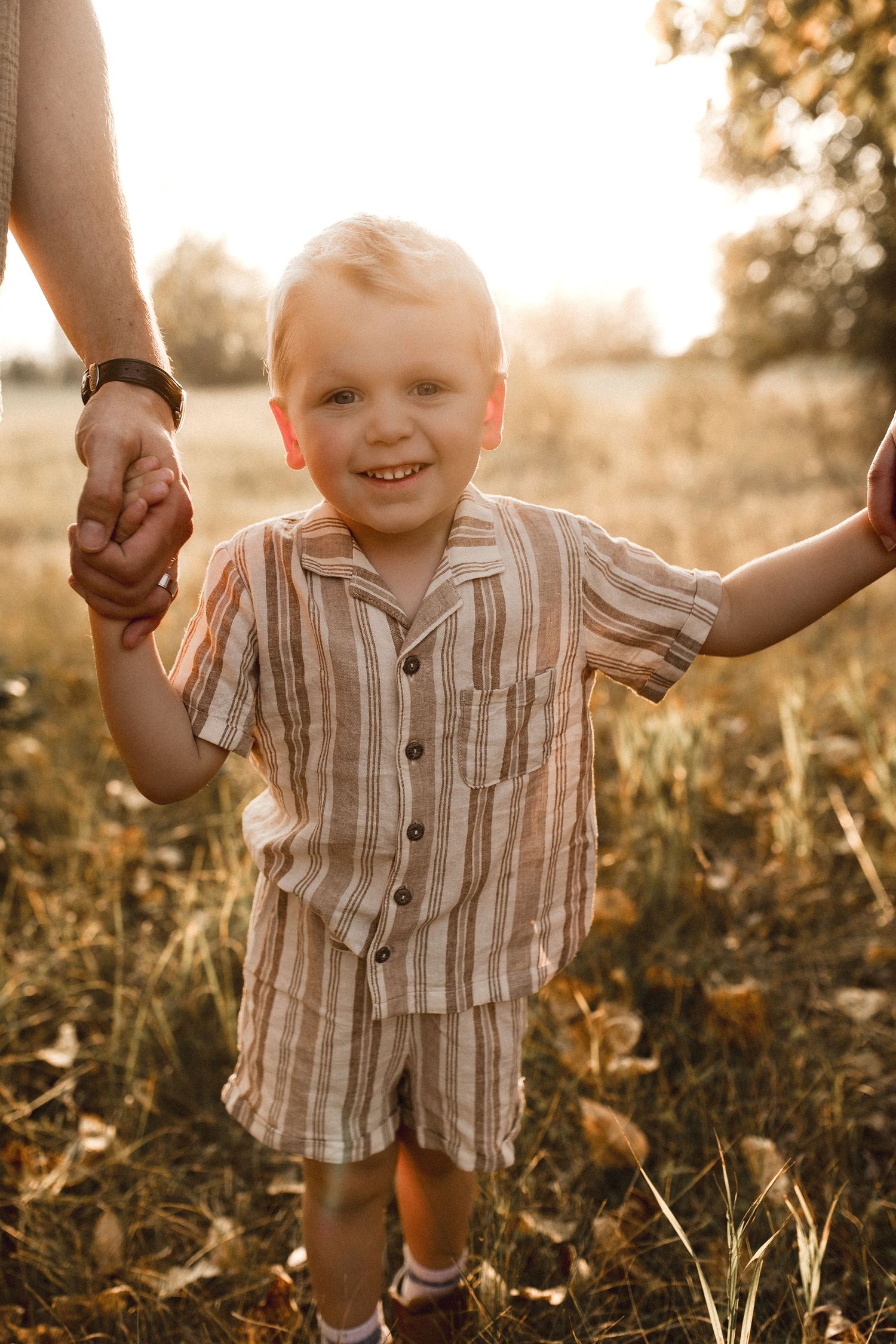 A happy toddler boy in striped outfit holds hands with adults while walking in a sunny golden field.