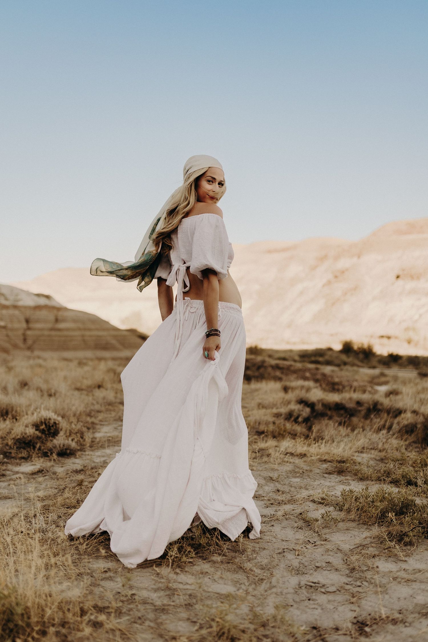 Woman in white boho crop top and flowing skirt twirling in a desert landscape at golden hour.