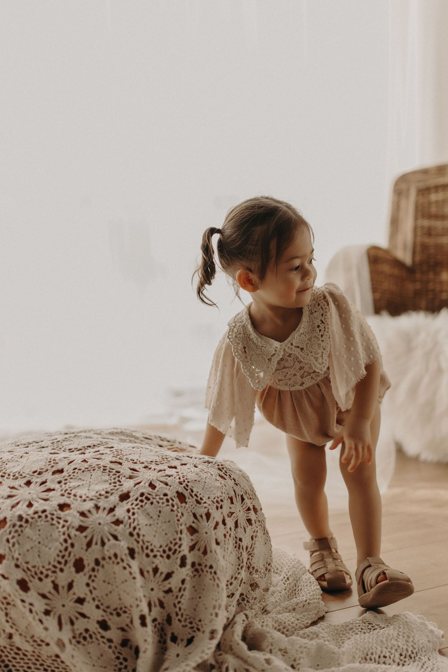 Young girl in crochet collar romper and sandals playing near a decorative blanket in a bright, airy room.