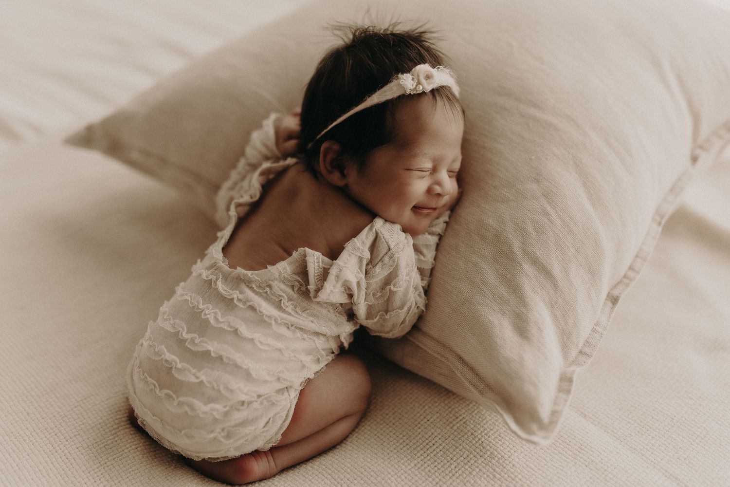 Peaceful newborn baby sleeping on a linen pillow, wearing a white lace romper and floral headband.