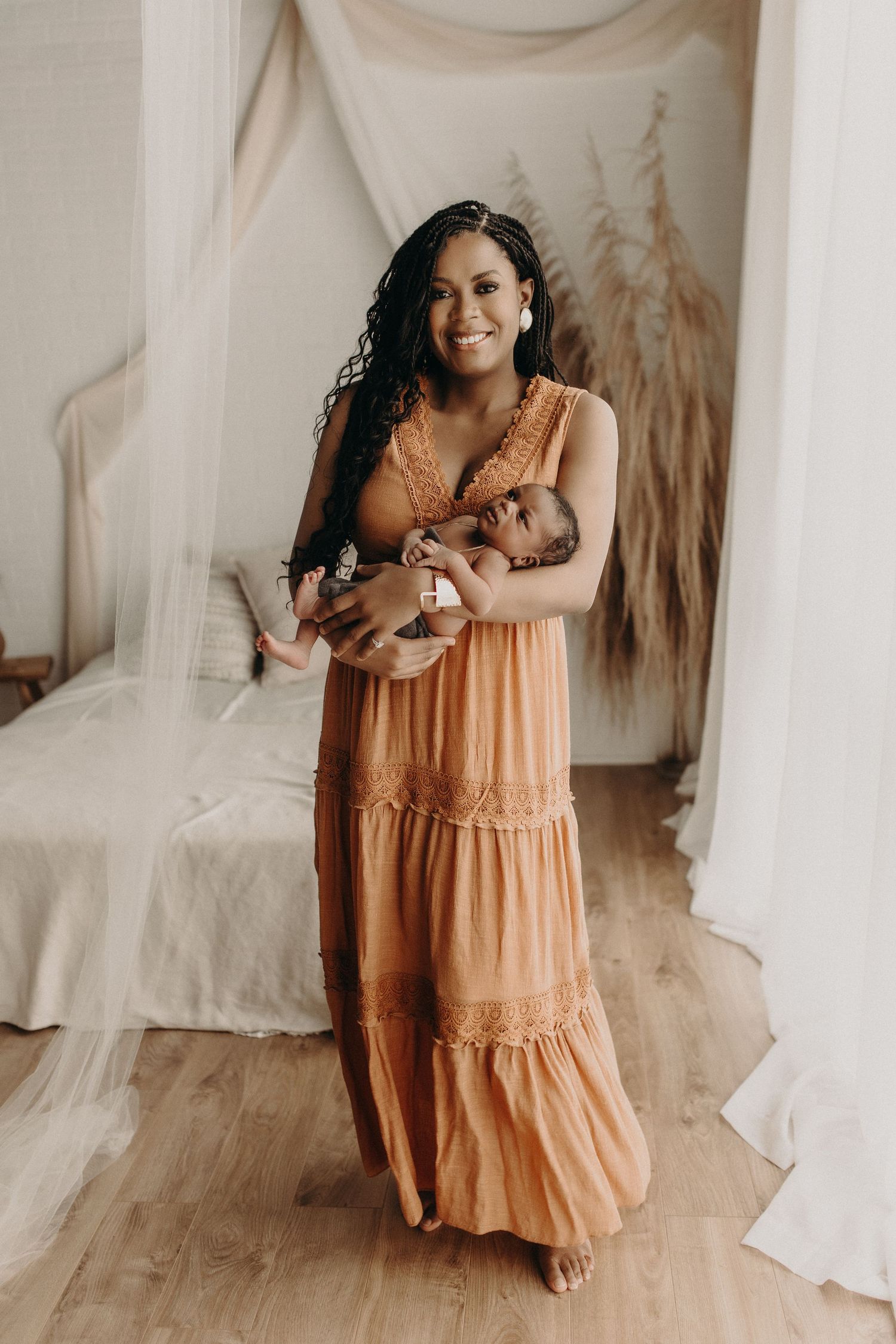 Smiling woman in orange boho maxi dress holding newborn baby in a softly decorated bedroom.