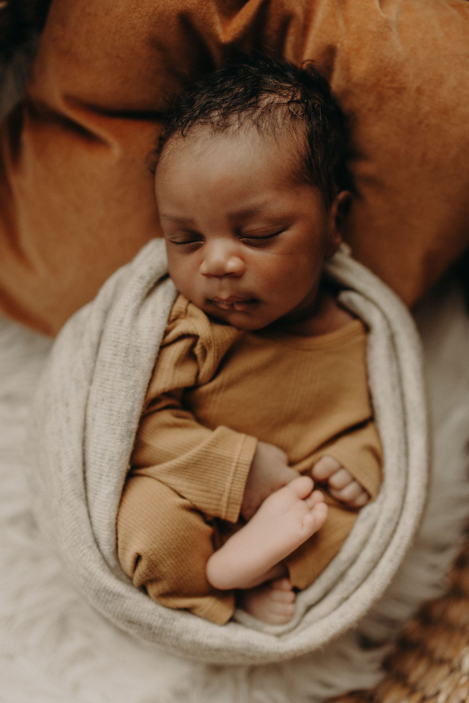 Newborn baby sleeping peacefully wrapped in a soft gray blanket wearing a mustard onesie nestled on a fluffy white surface.