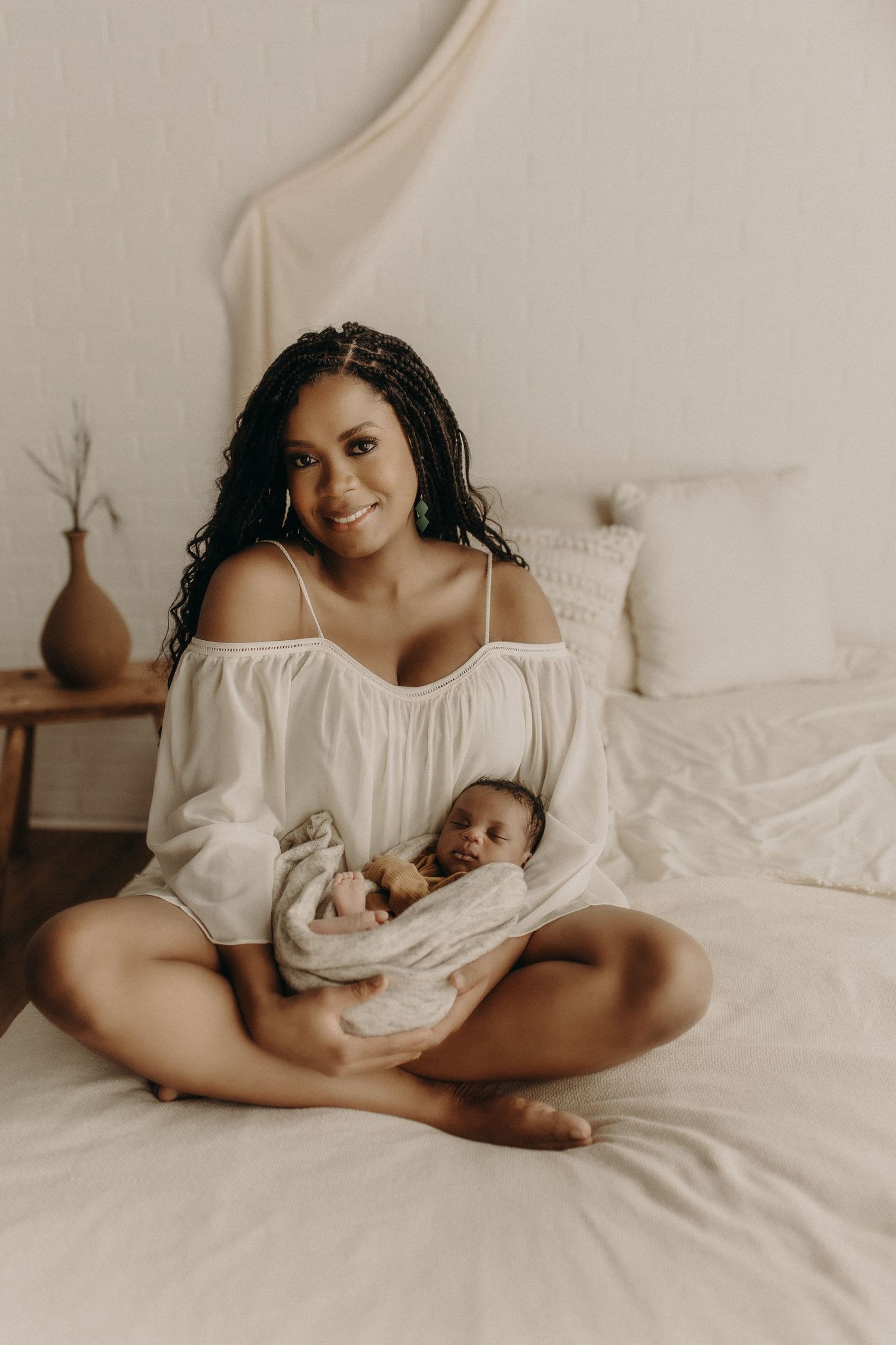 A smiling woman in a white off-shoulder dress sits cross-legged on a bed, cradling a swaddled newborn baby.