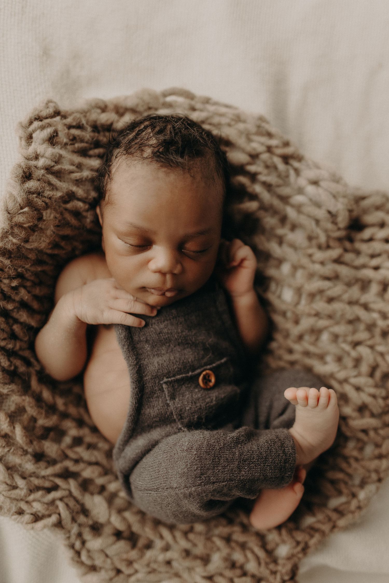 Newborn baby sleeping peacefully in a knit basket wearing gray overalls during a professional newborn photo session.