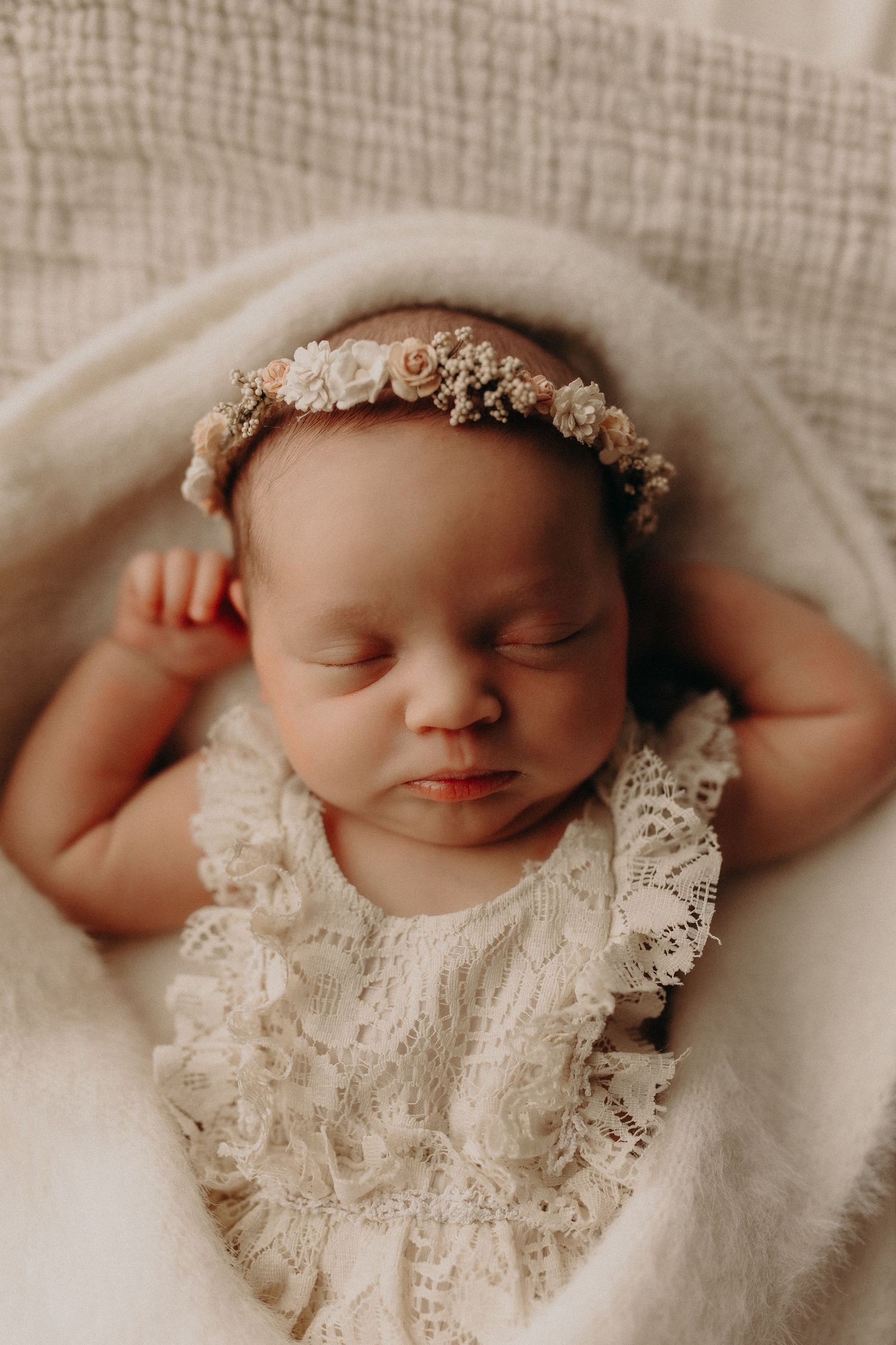 Sleeping newborn baby girl wearing a white lace romper and delicate floral headband, nestled in a soft cream blanket.