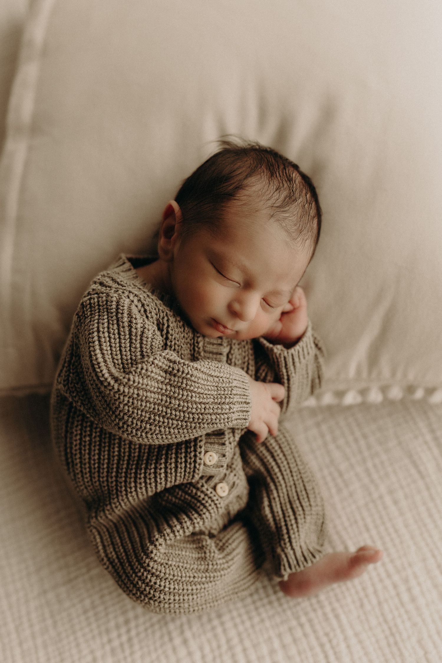 Sleeping newborn baby in a chunky knit olive romper, peacefully resting on a cream textured surface.