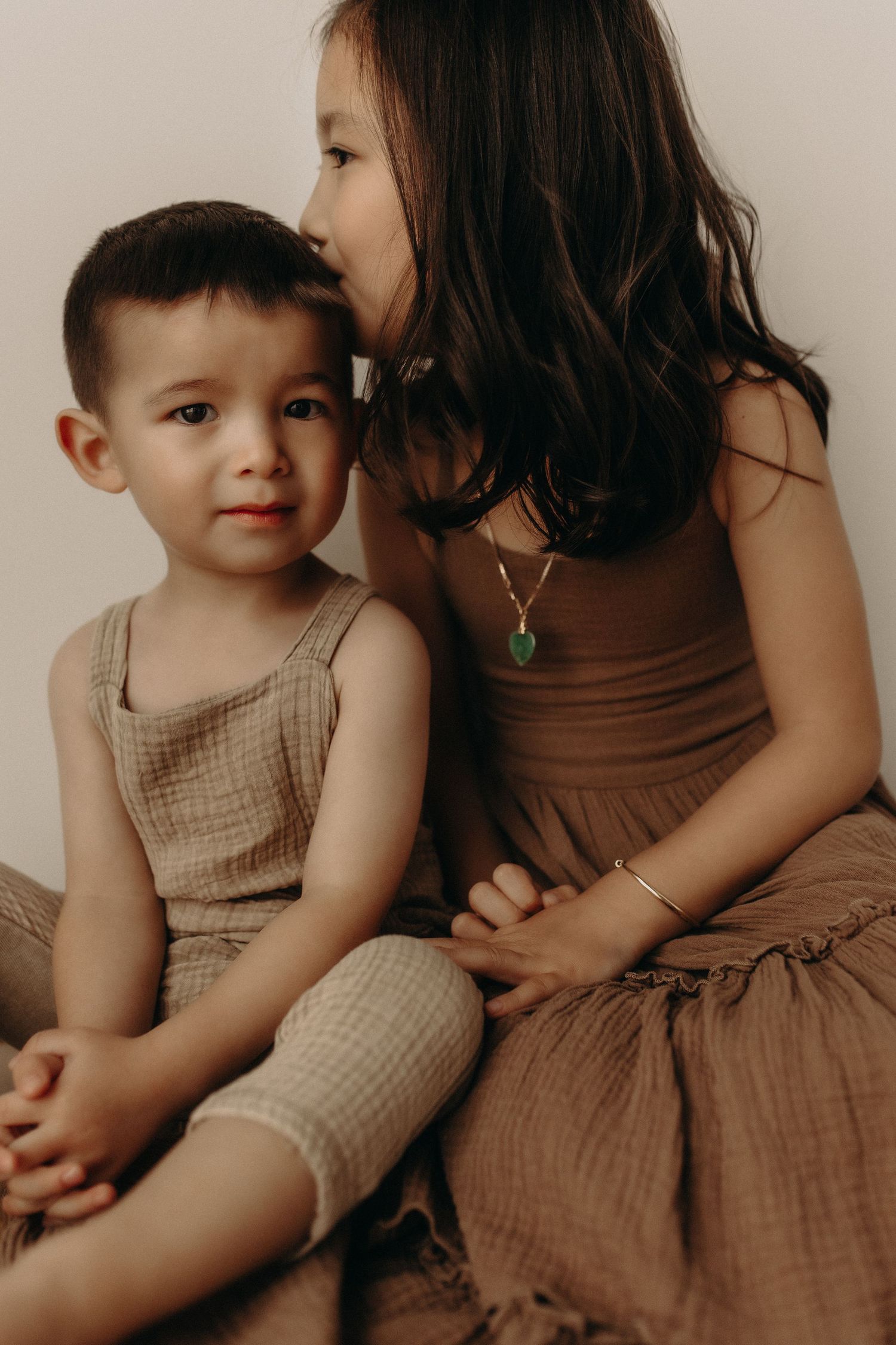 Two young siblings in earthy tones, older girl kissing younger boy on cheek, wearing linen outfits and gold jewelry.