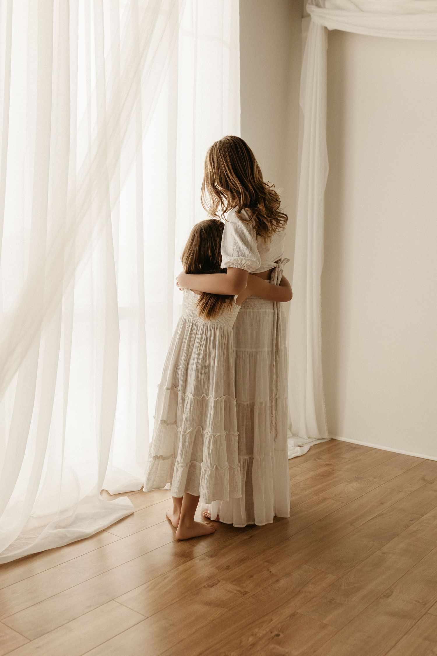 A mother and child embrace while gazing out a bright window, both wearing flowing white and beige dresses on hardwood floors.