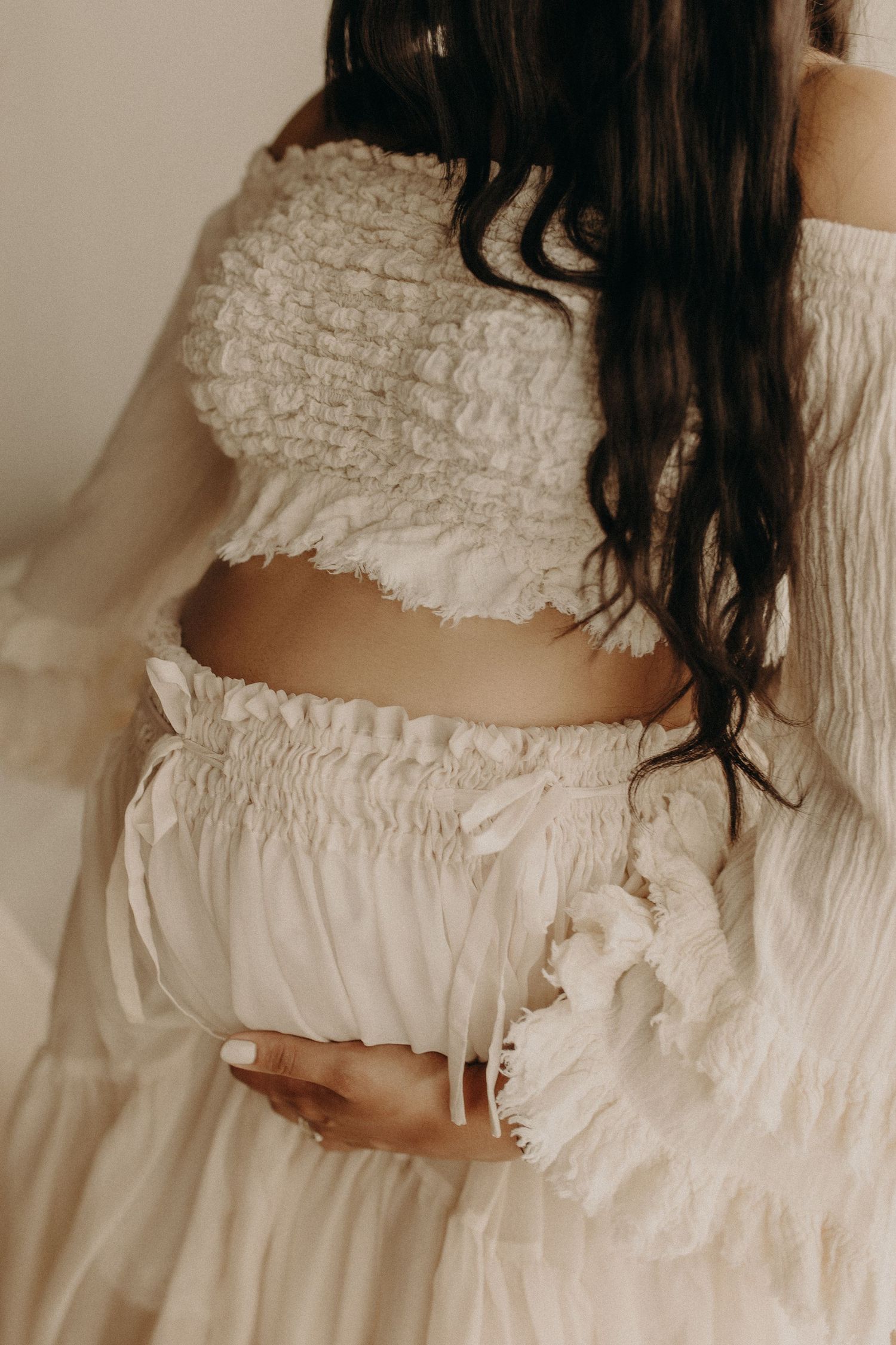 Pregnant woman in white ruffled crop top and skirt cradling baby bump, boho maternity photoshoot.