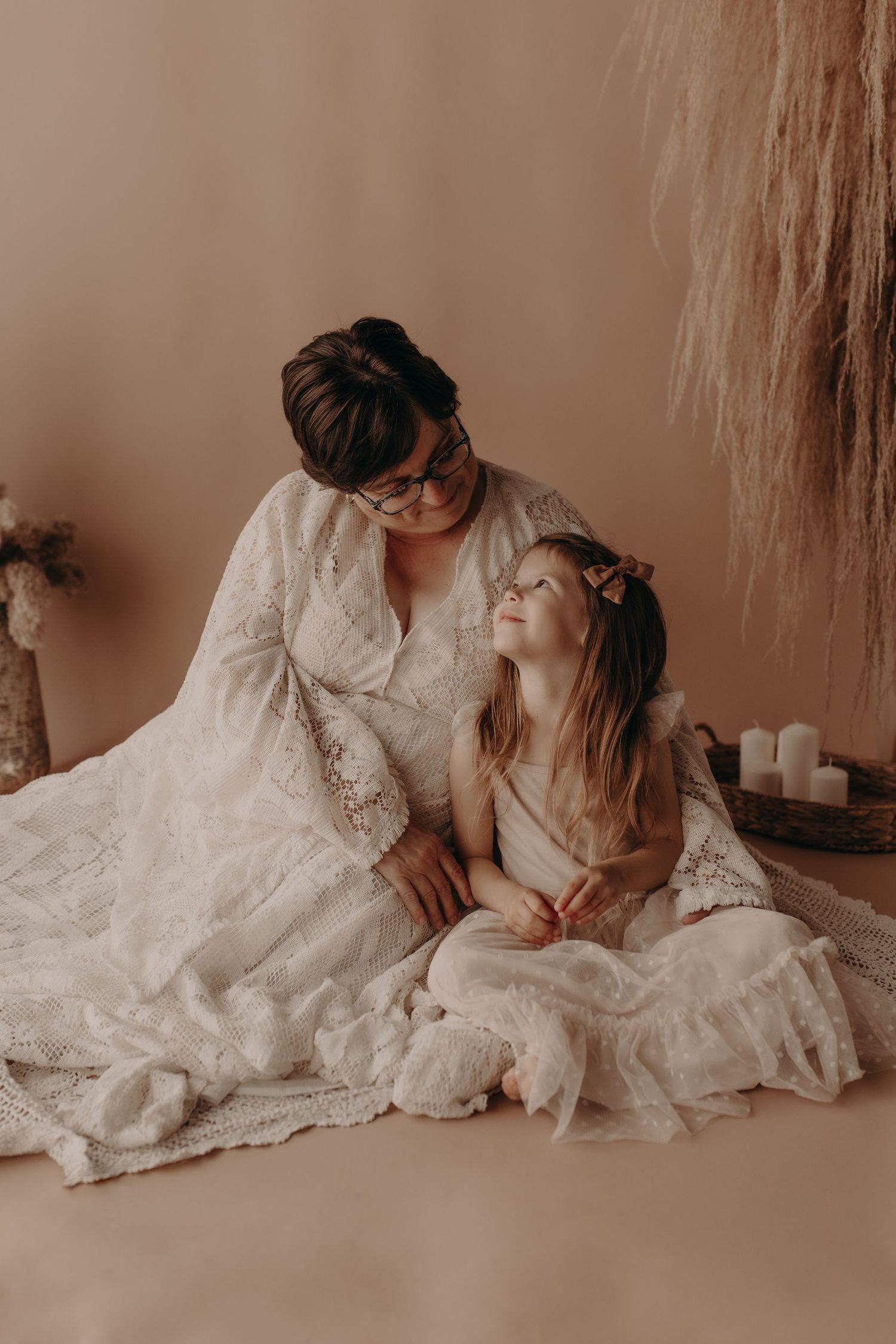 A woman in a white lace dress sits on the floor with a young girl in a white dress, sharing a tender moment.
