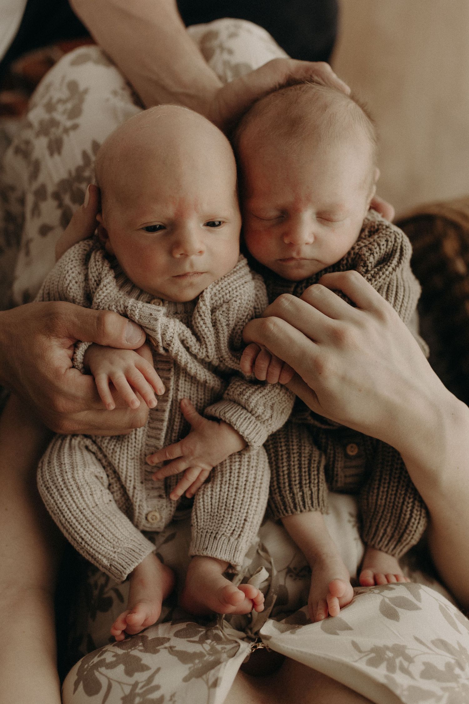 Two newborn twins in matching knit onesies being held by their parent, with tiny feet and hands visible.