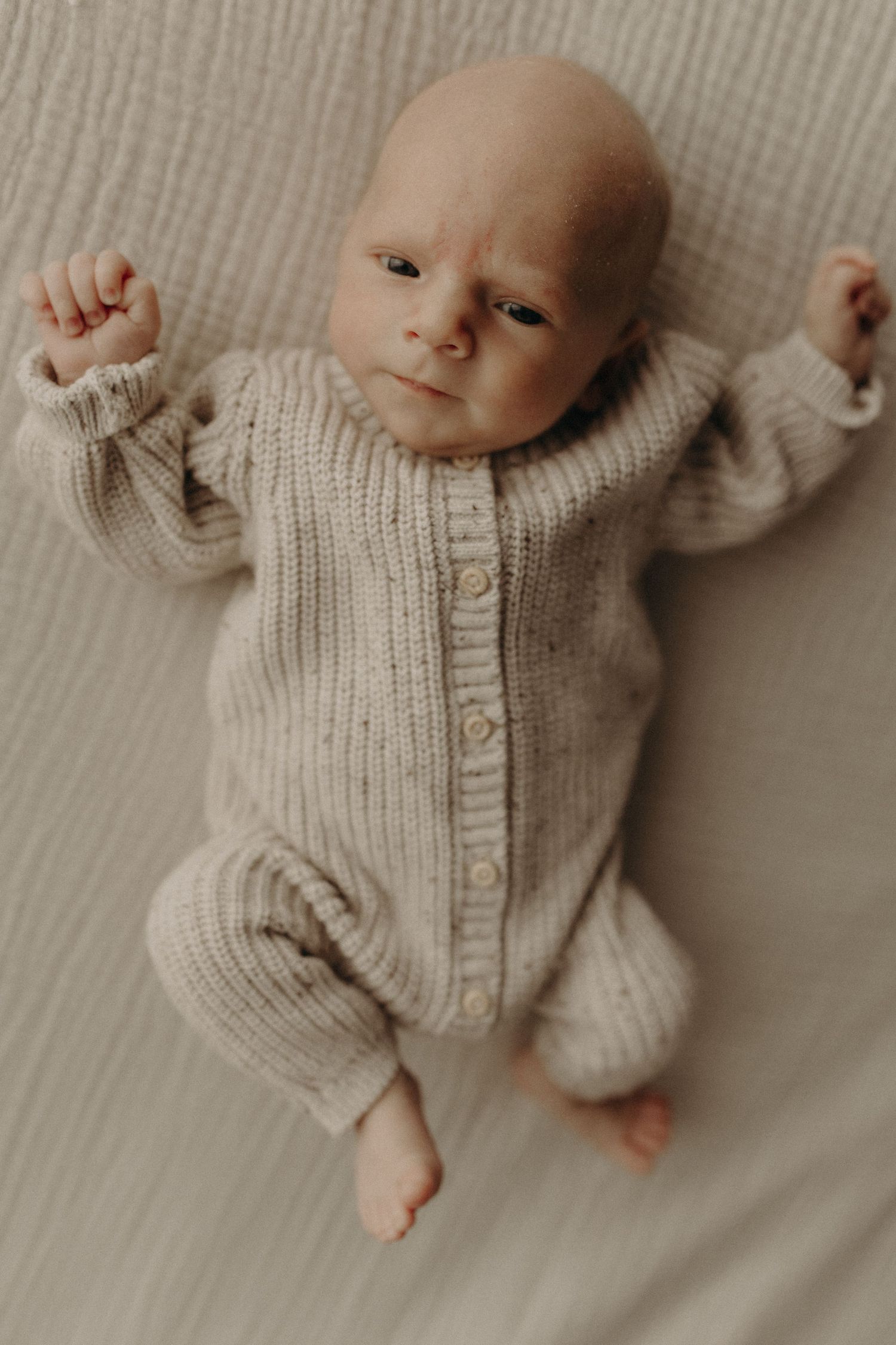 Newborn baby in a beige knit romper lying on a white blanket, arms raised, looking up with wide eyes.