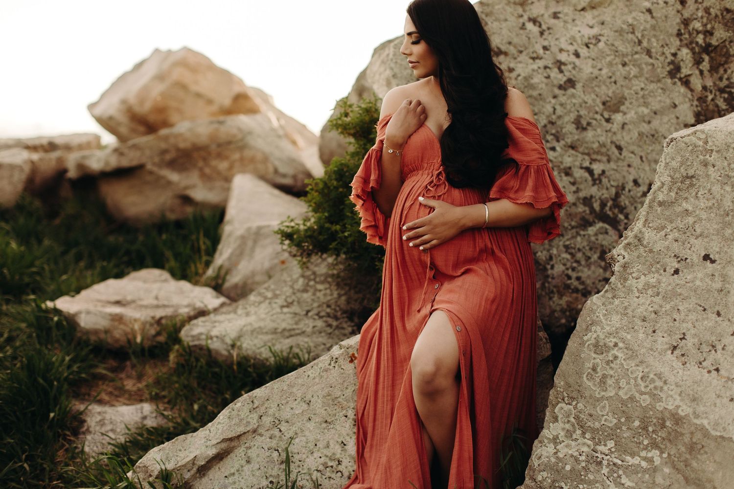 Pregnant woman in flowing rust-orange maternity gown poses gracefully among large rocks in an outdoor setting.