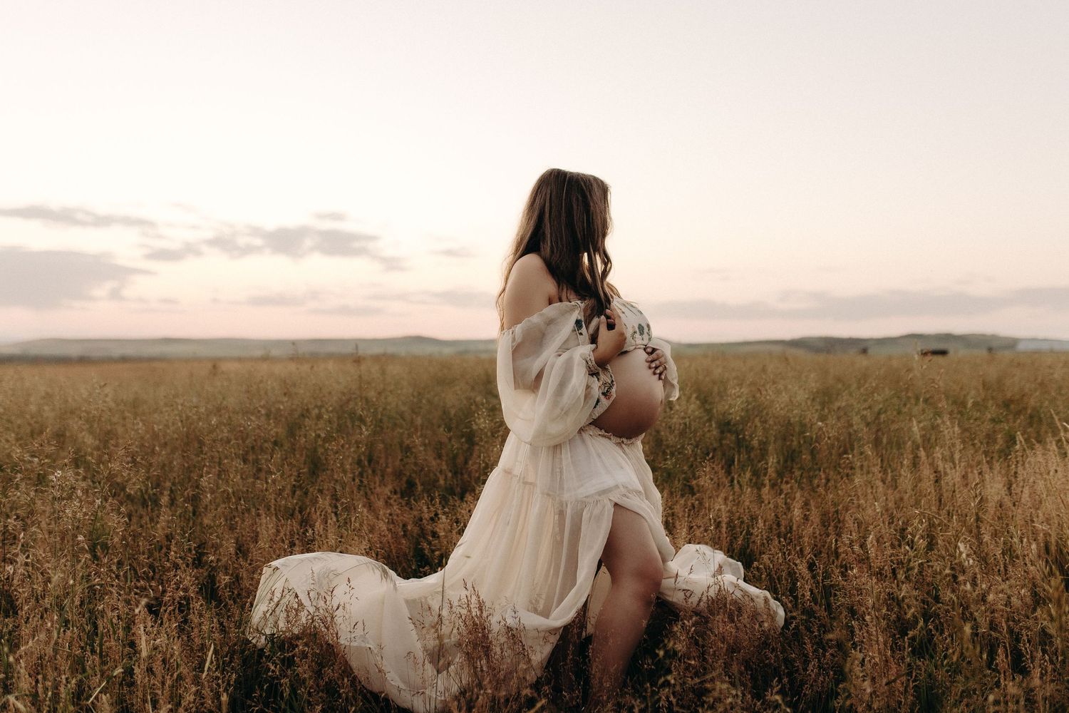Pregnant woman in white dress standing in golden field at sunset, cradling her baby bump in a serene outdoor maternity photo.