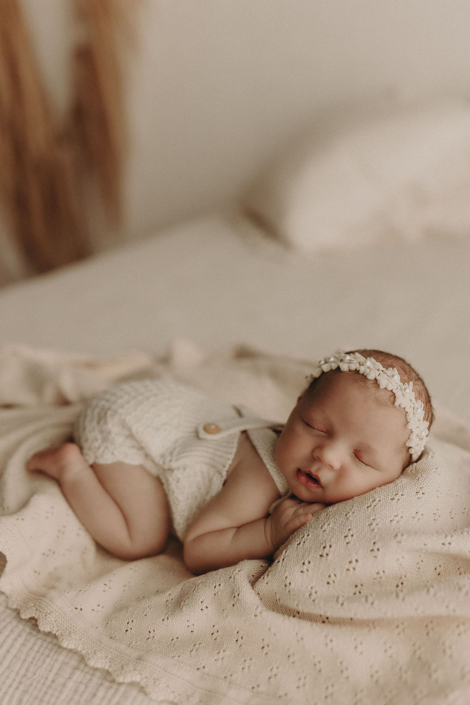 Sleeping newborn baby girl in cream knit romper and lace headband on soft beige blanket.