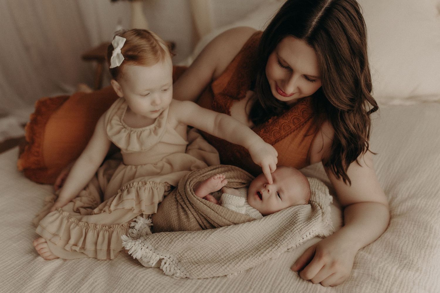 A mother smiles lovingly at her newborn baby while her toddler daughter gently touches the infant on a bed.