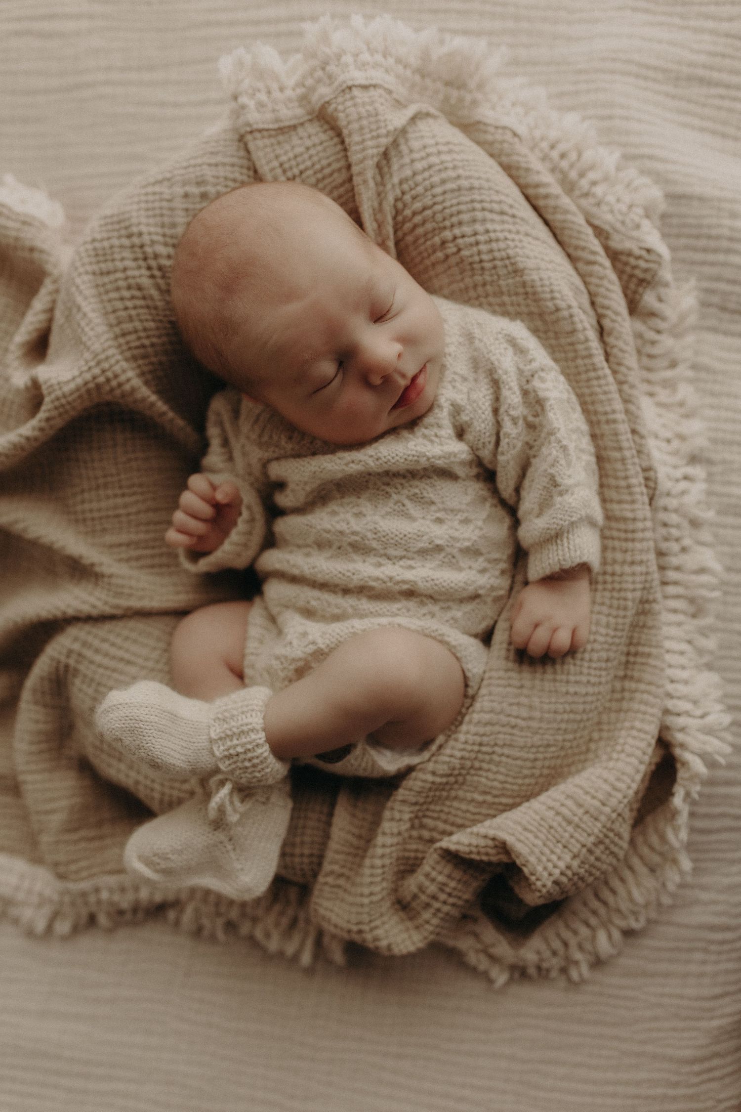 A peaceful newborn baby sleeps wrapped in a cozy beige knit outfit and blanket with tiny white socks.