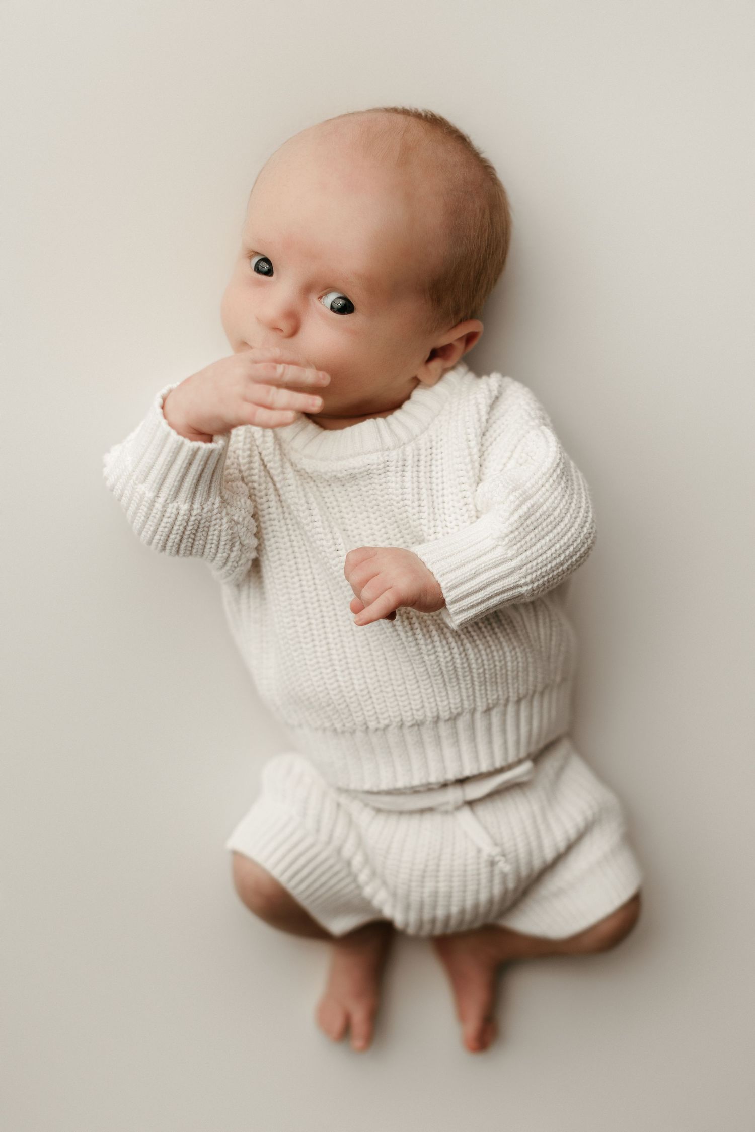 Newborn baby in white knit sweater and shorts lying on a light background, gazing up with hand near mouth.