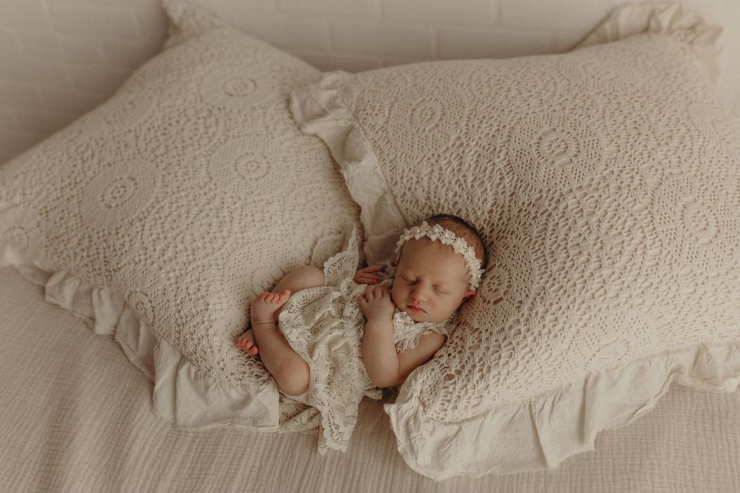 Newborn baby sleeping peacefully between two cream ruffled pillows wearing a lace romper and floral headband.