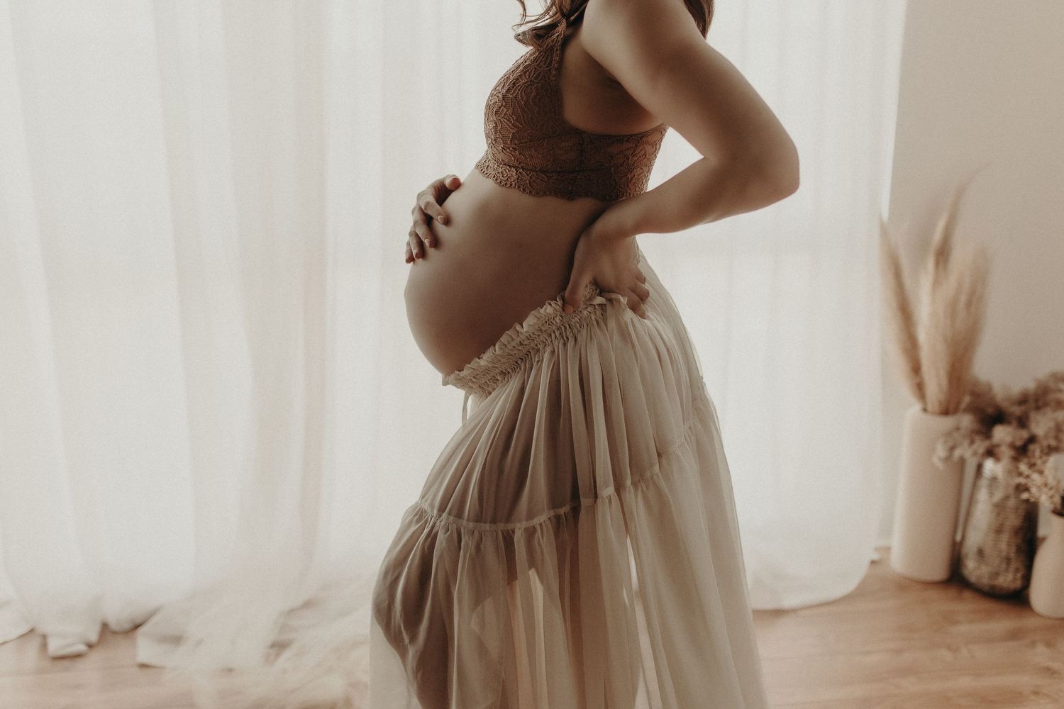 Pregnant woman in brown lace bralette and flowing tulle skirt cradling her baby bump in a softly lit room.
