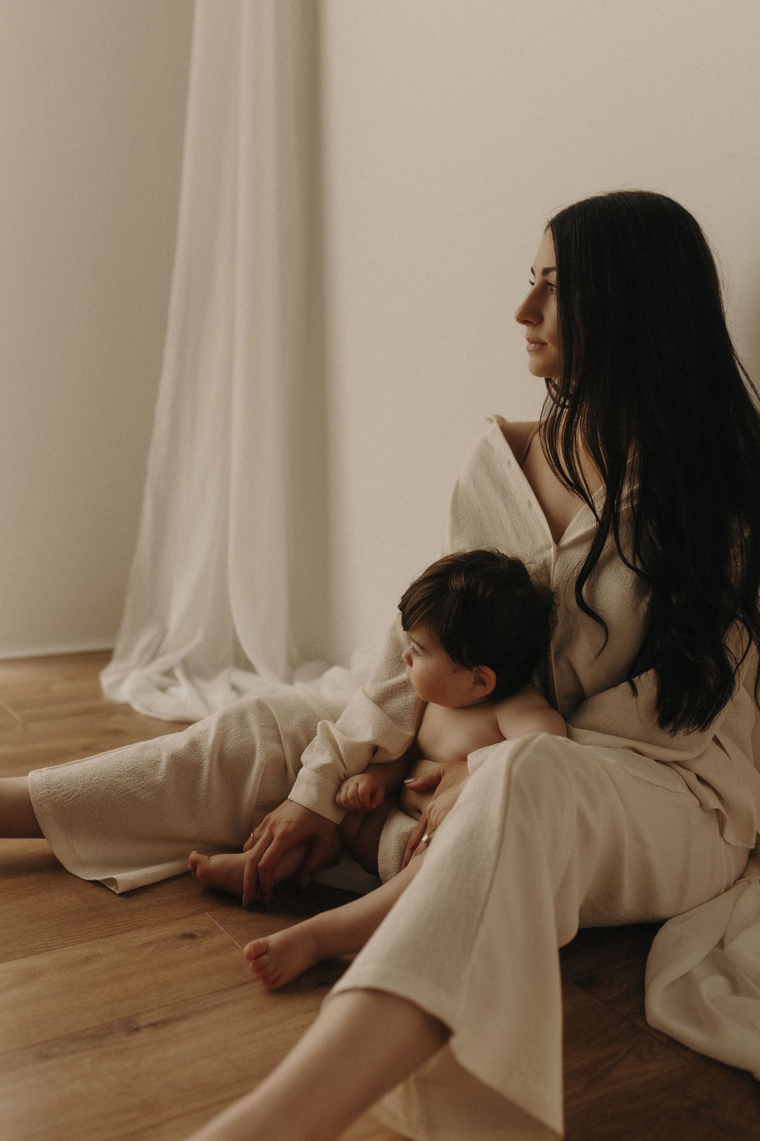 A mother breastfeeding her toddler while sitting on a wooden floor in soft white natural light.