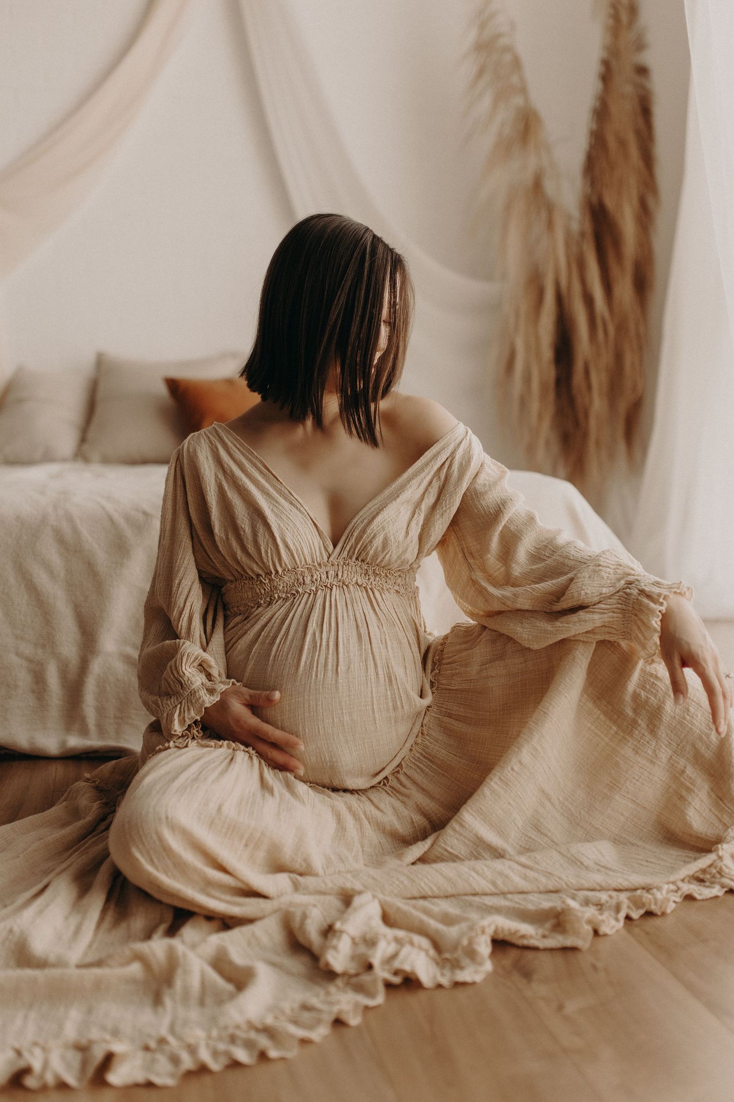 Pregnant woman in beige linen dress sitting on floor, cradling baby bump in boho bedroom setting.